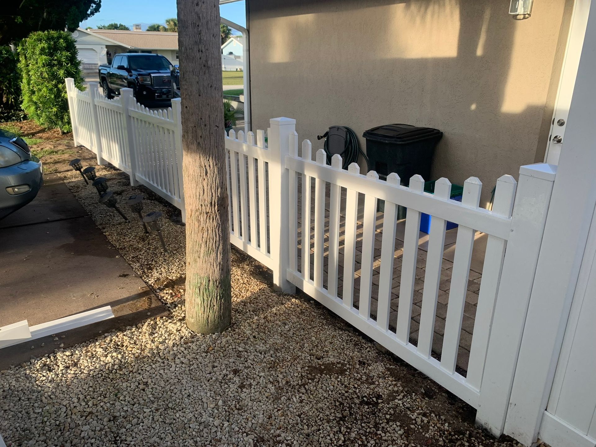 White picket fence along a gravel driveway beside a house, with a tree and parked cars nearby