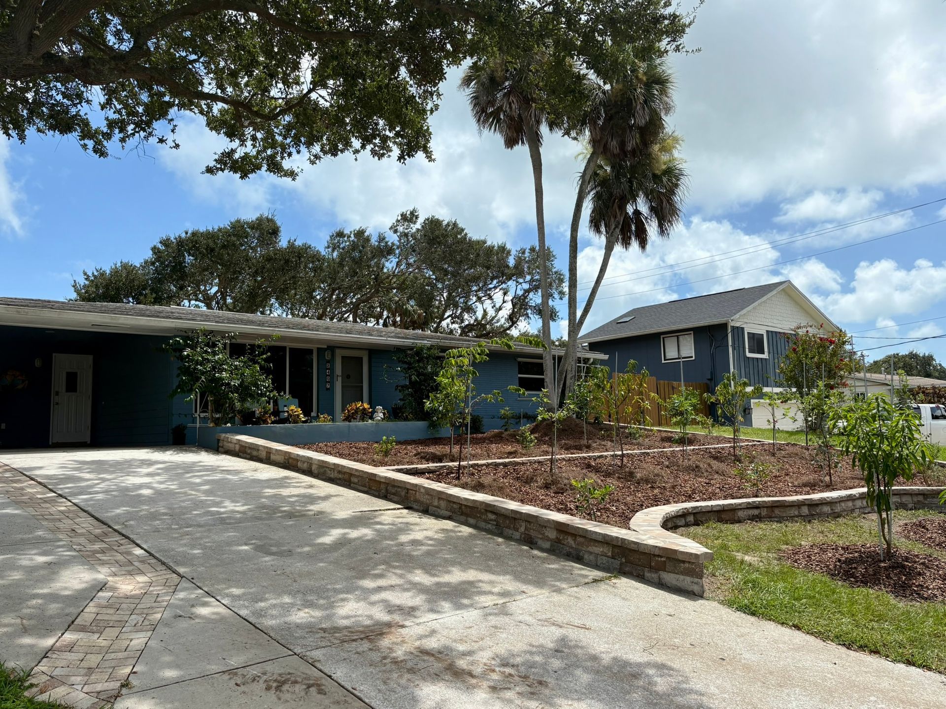 Blue house with driveway, palm trees, and landscaped front yard under a partly cloudy sky