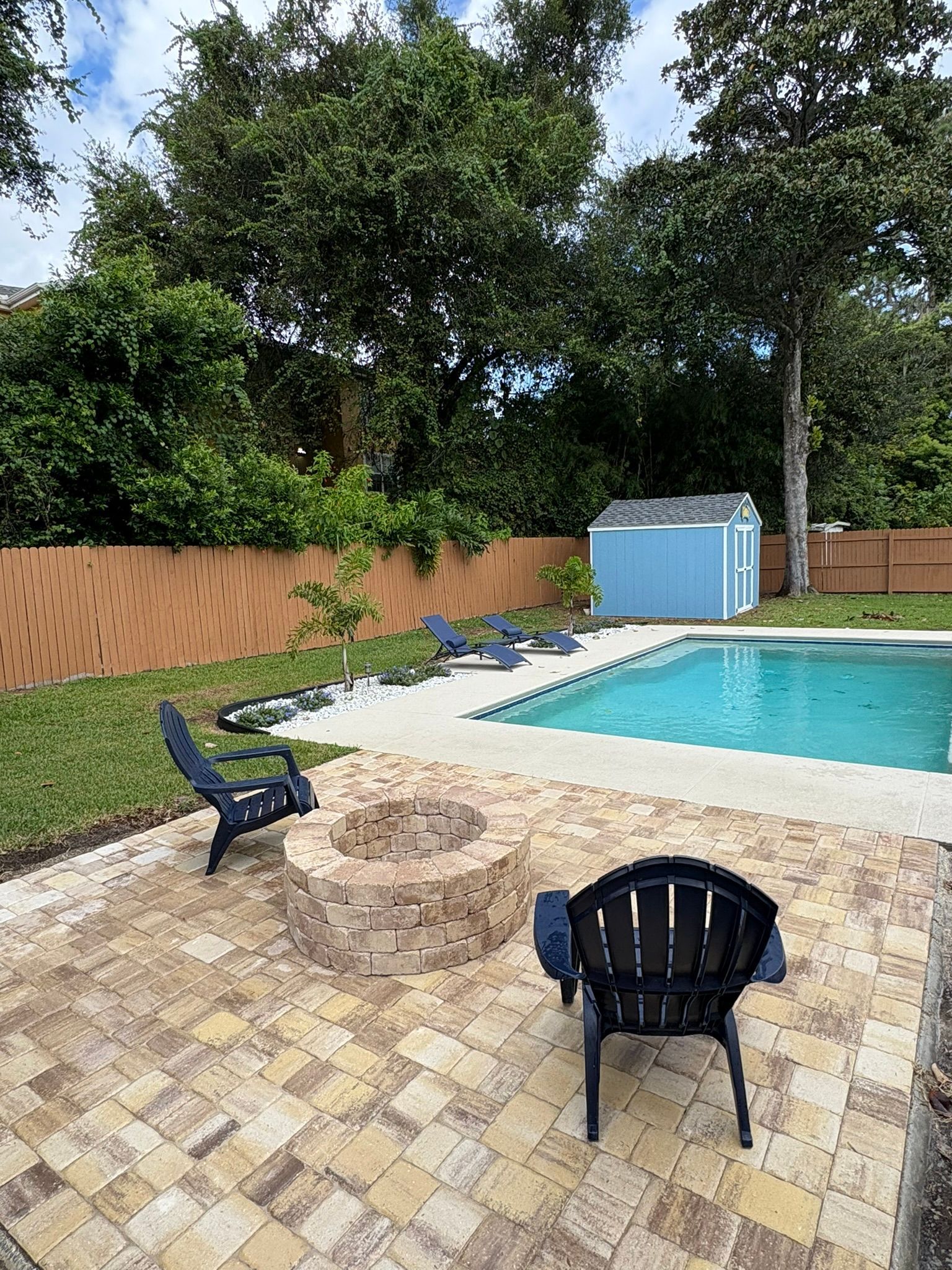 Backyard pool with blue water, lounge chairs, tan patio tiles, and a blue storage shed surrounded by trees