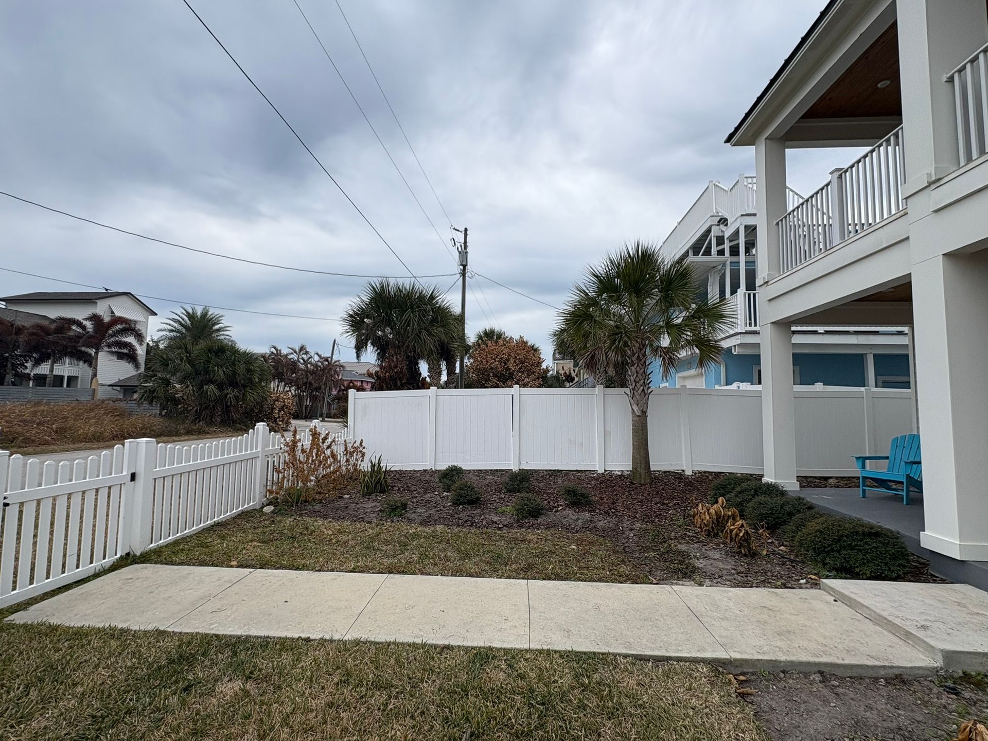 Small front yard with white fence, sidewalk, palm trees, and a house on a cloudy day