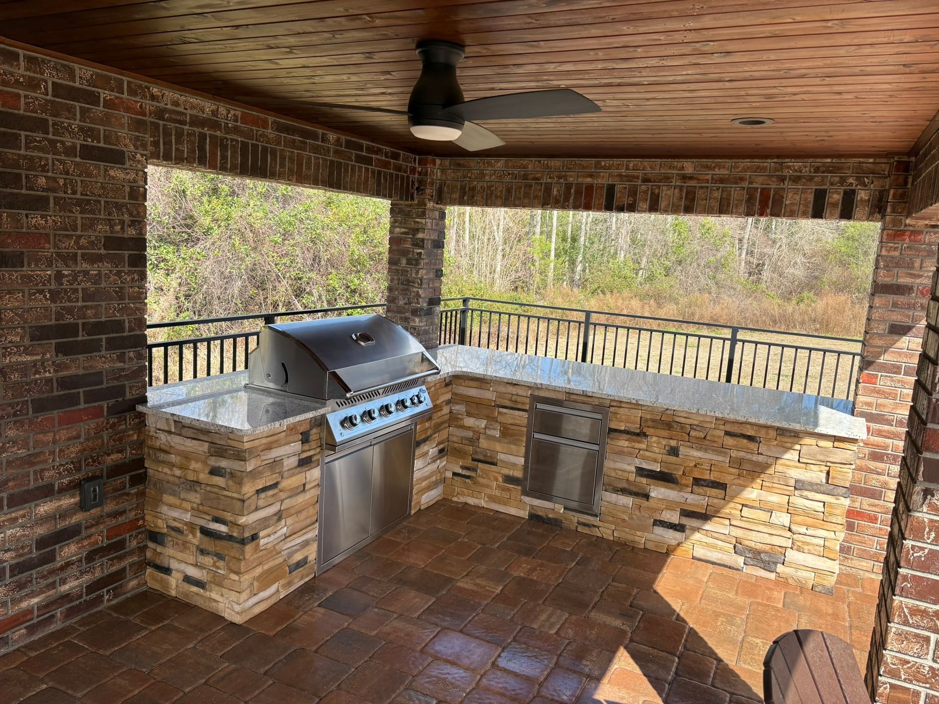 Covered outdoor kitchen with brick grill, stainless steel sink, and tiled floor overlooking the woods