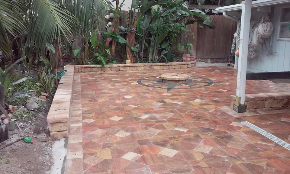 Brick patio with a central decorative element, surrounded by greenery and a wooden fence.