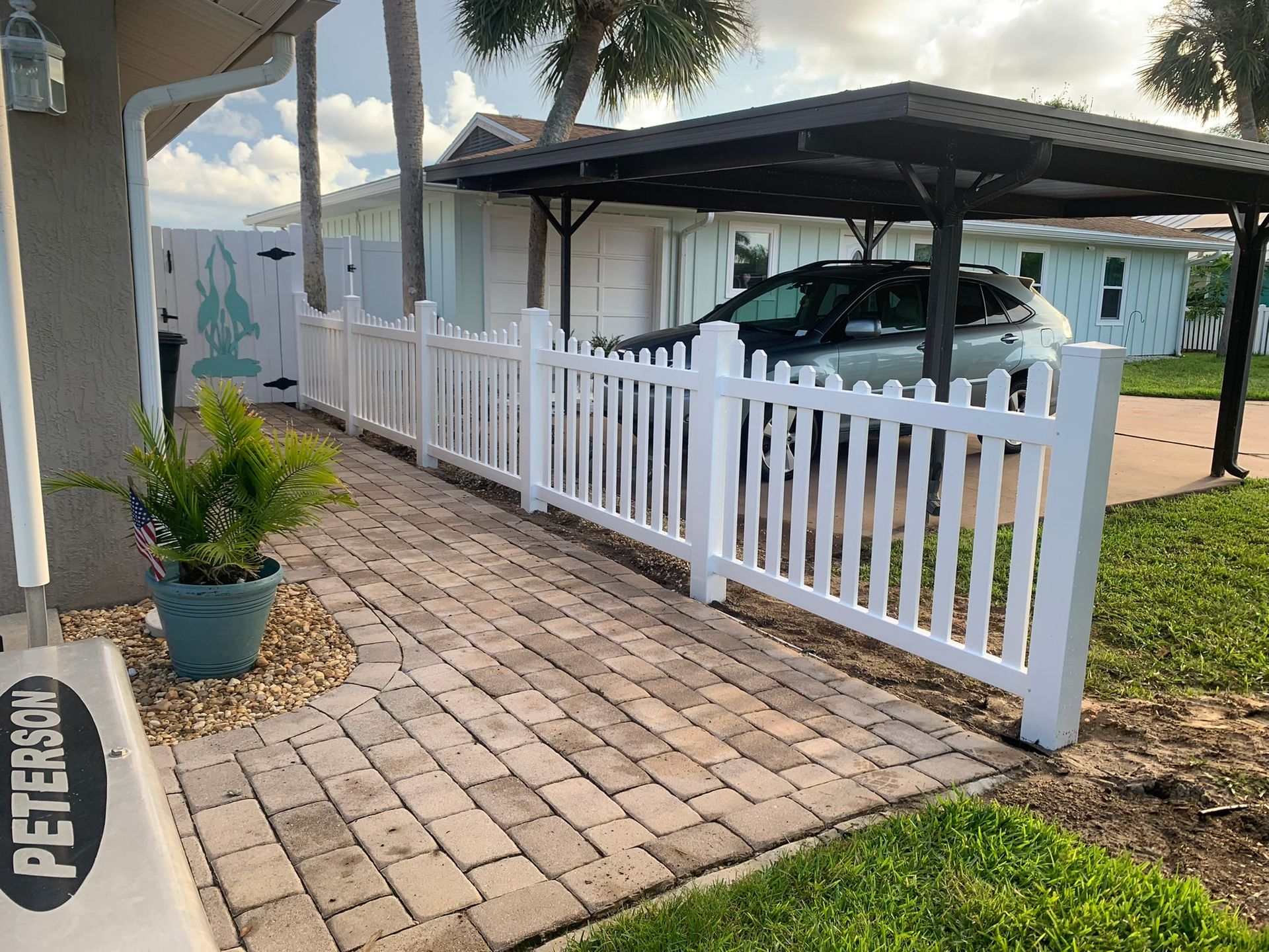 White picket-fenced carport with a silver car, paver walkway, and potted plants beside a blue house.