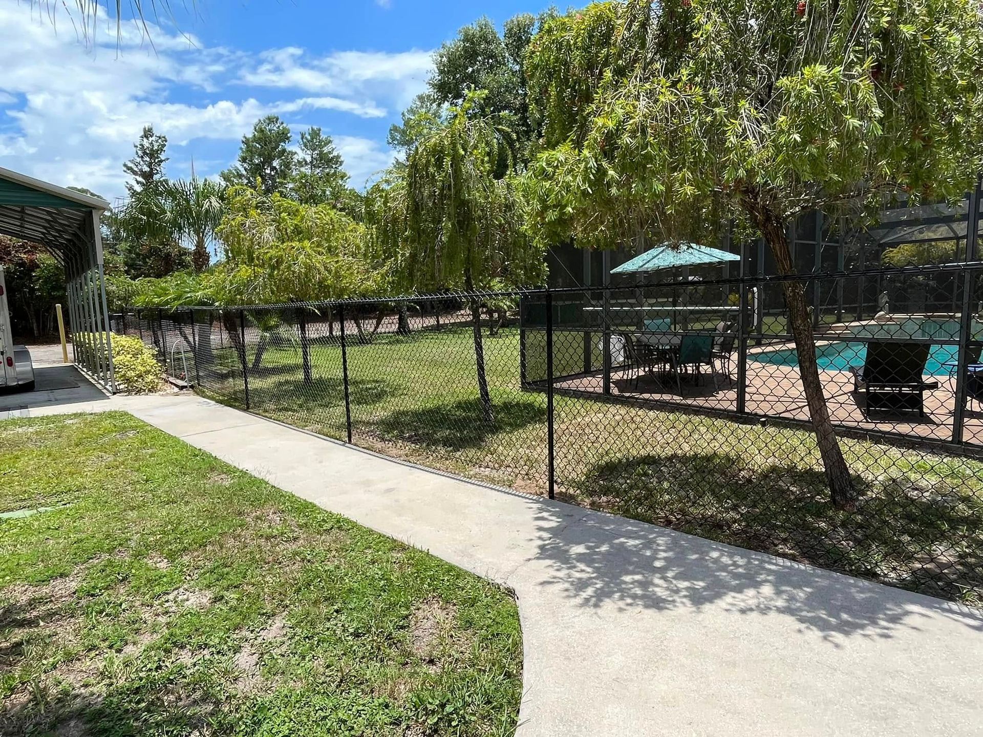 A concrete path alongside a chain-link fence, leading to a pool area. Green trees and a blue sky are visible.
