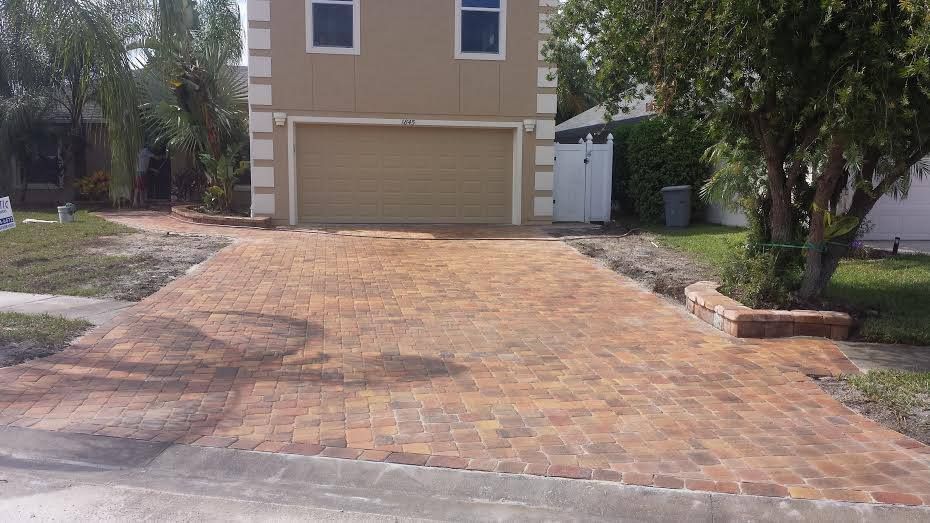 Brick driveway leading to a two-story beige house with a garage.