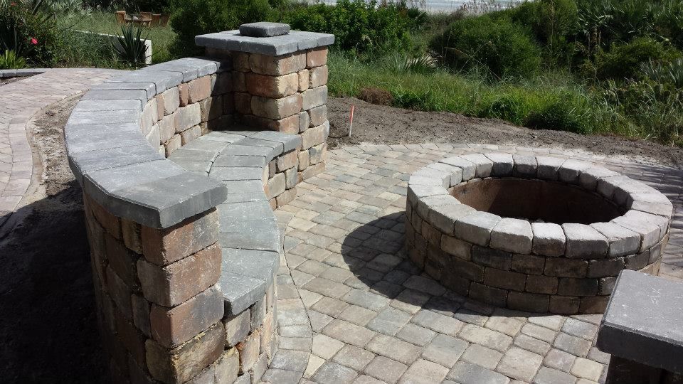 Brick-built outdoor seating area and fire pit on a paved patio with green grass and foliage in the background.