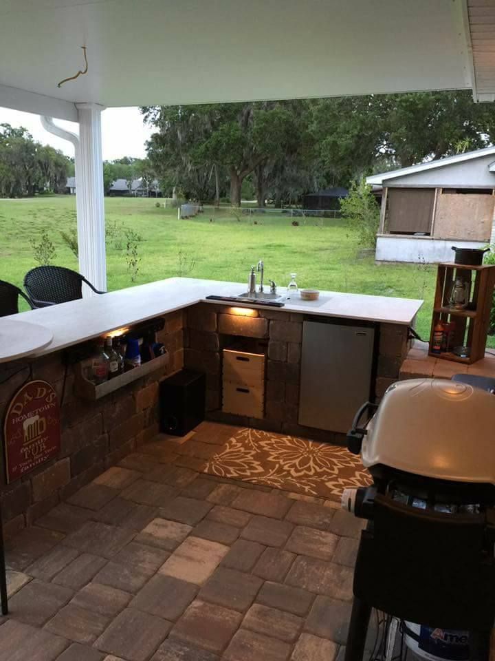 Outdoor kitchen with stone facade, concrete countertop, sink, fridge, grill, and bar seating.