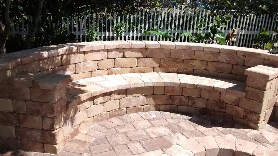 Stone bench in a circular patio, built of red-brown bricks. White picket fence and greenery in the background.