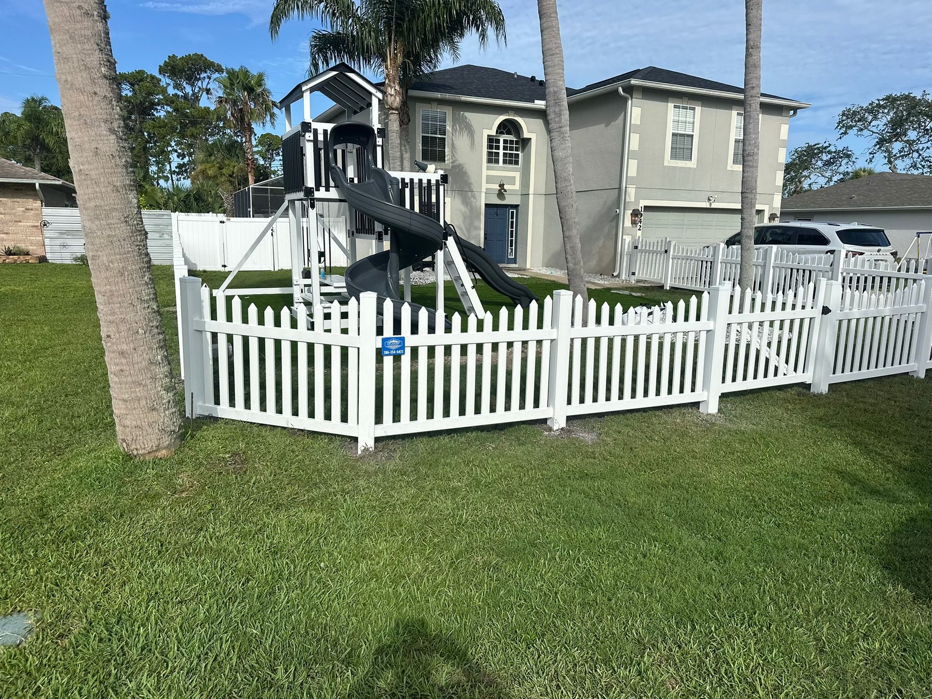 White picket fence surrounds a playground in a yard in front of a two-story house on a sunny day.