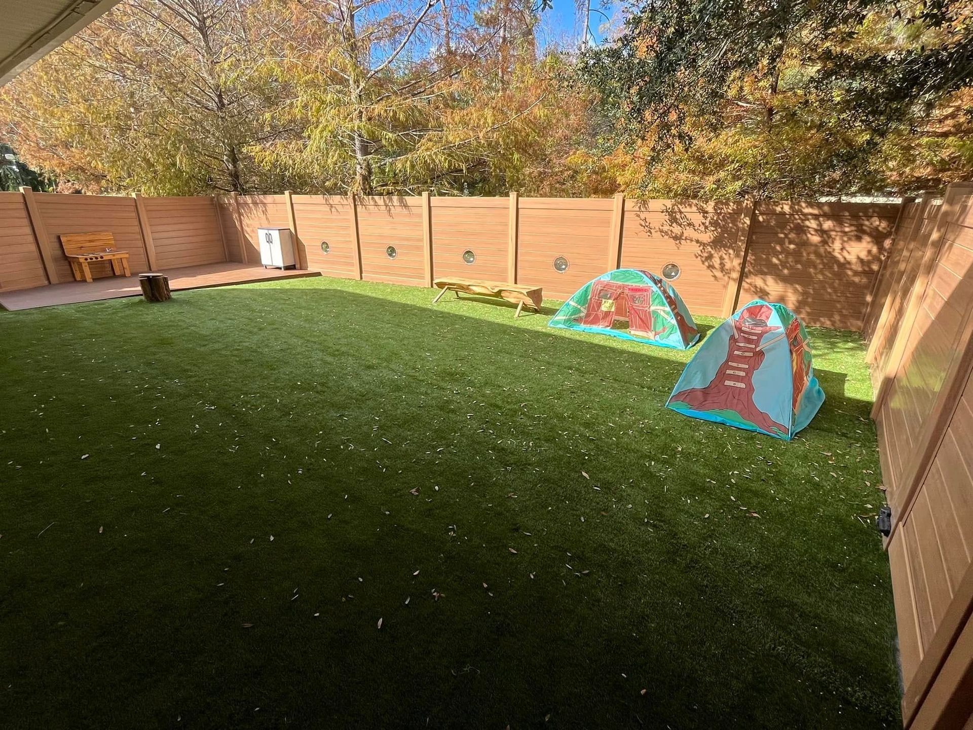 Backyard with artificial grass, two tents, wooden fence, and fall foliage.