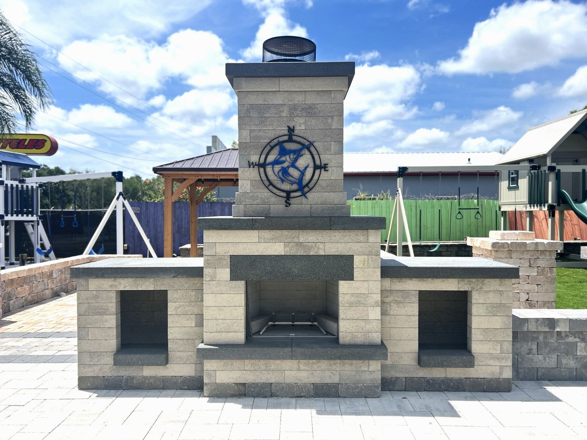 Outdoor stone fireplace with fish compass design, flanked by storage compartments, under a blue sky.