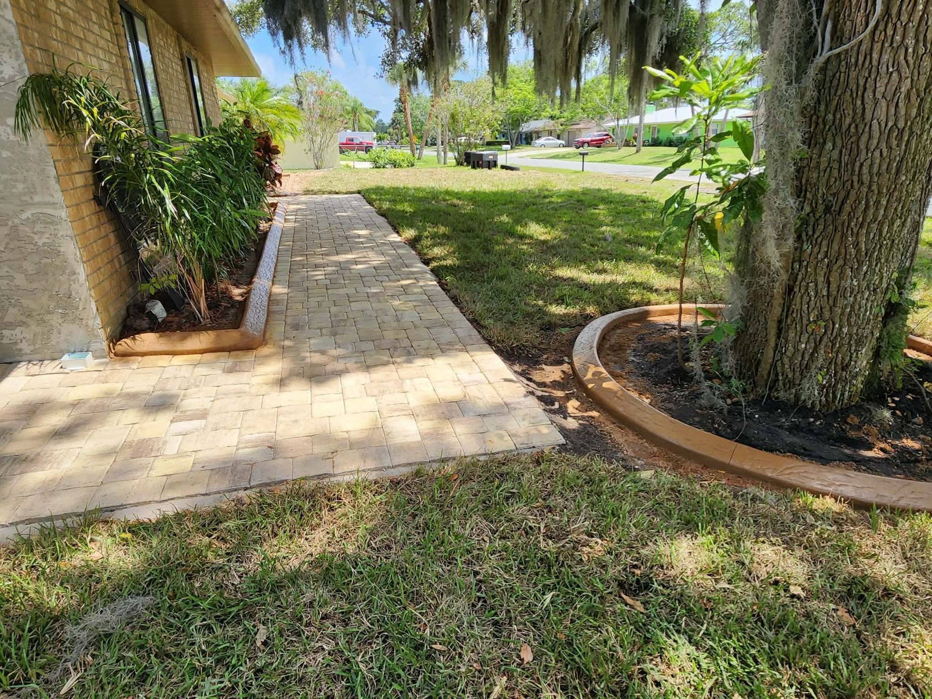 Brick walkway through a grassy yard, bordered by brown concrete, leading to a home.