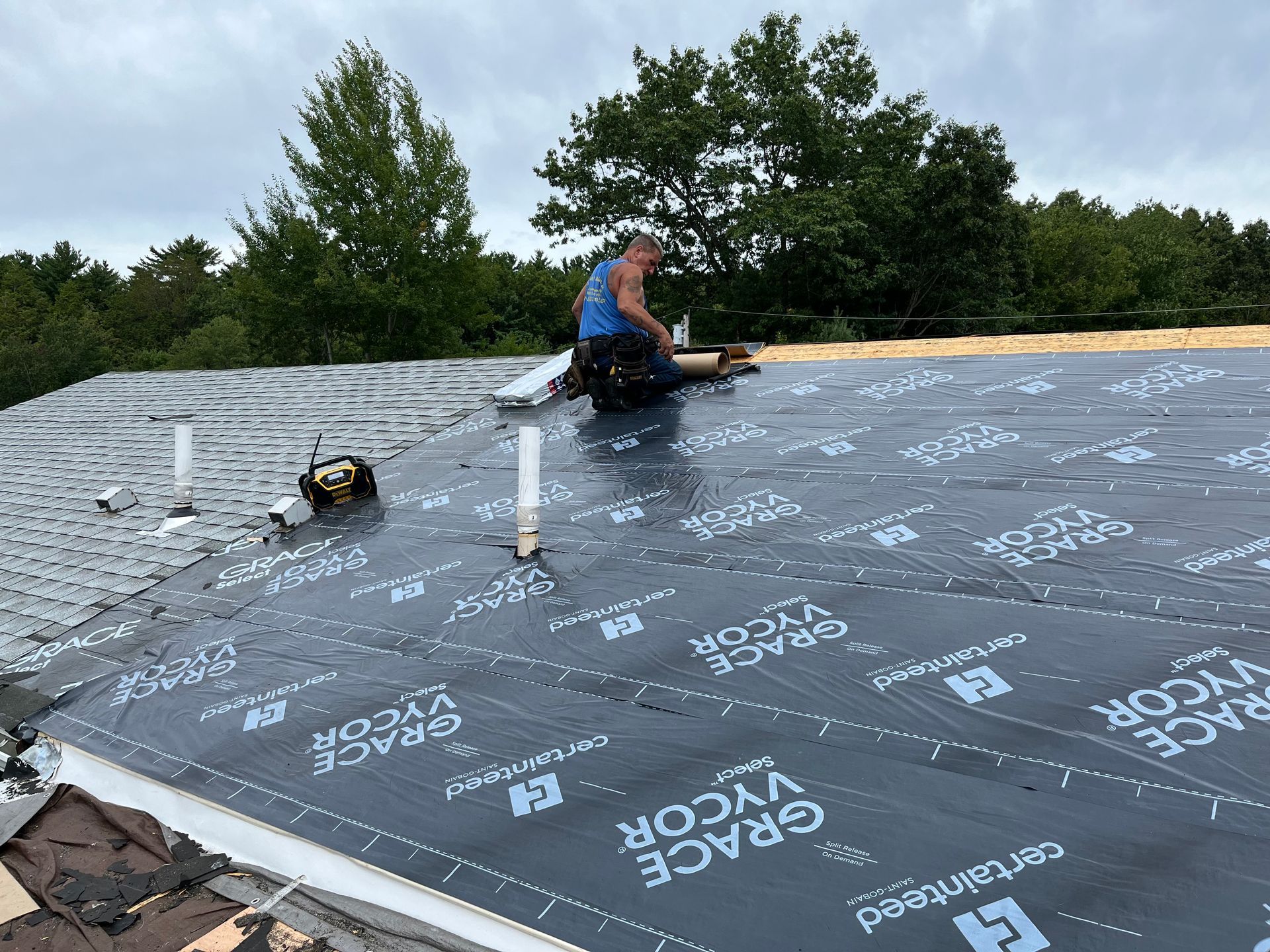 Worker kneels on a flat roof, installing black underlayment beside white vent pipes and trees in the background