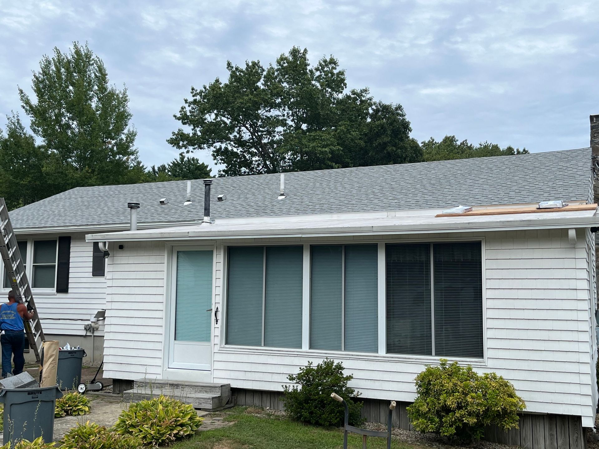 Single-story white house with a gray shingle roof and closed front windows under overcast skies