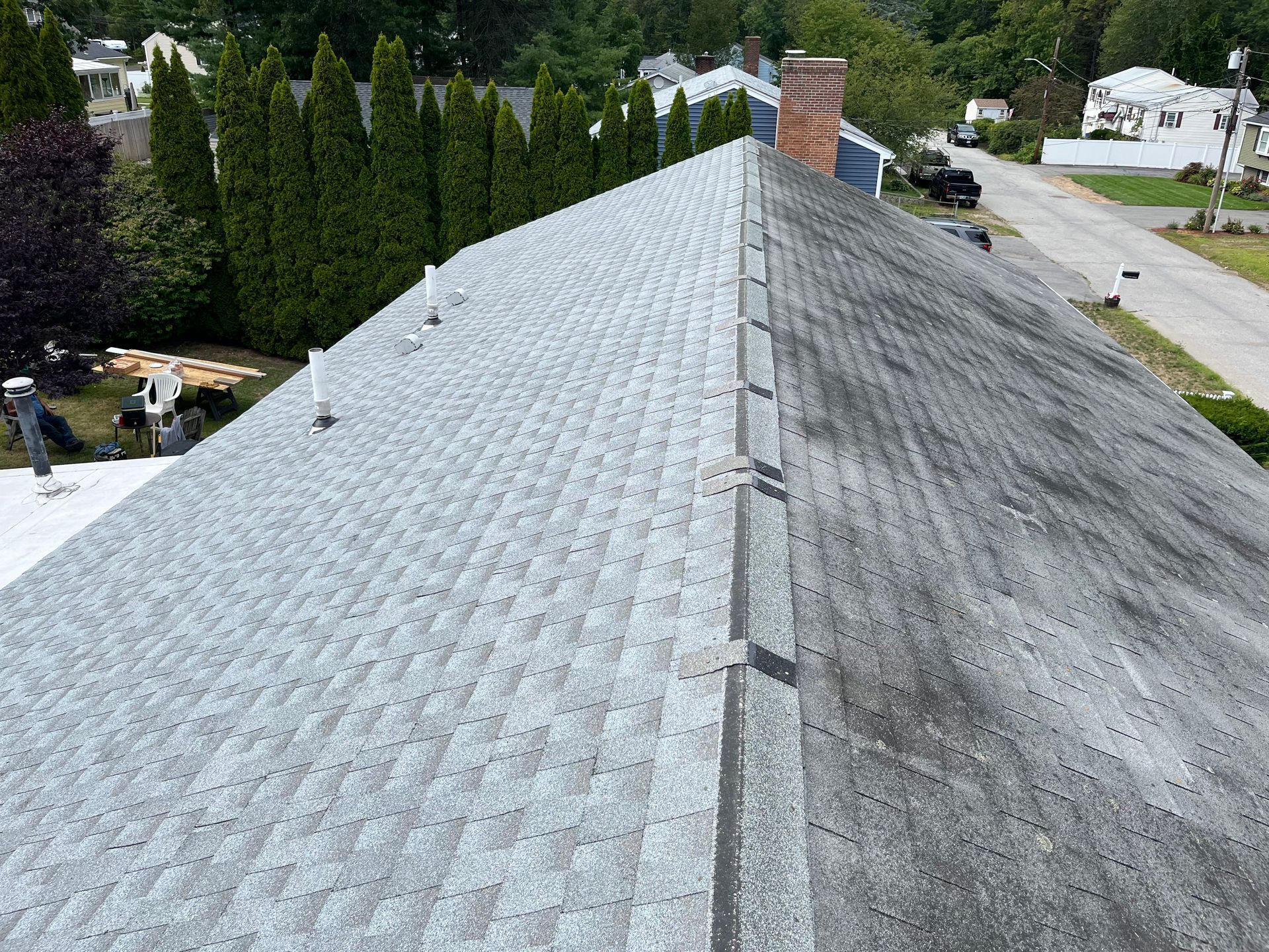 Aerial view of a gray shingled roof with a brick chimney and tall evergreens nearby.
