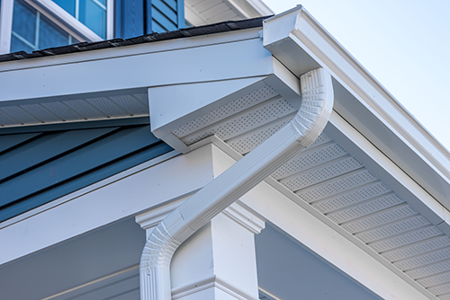 Close-up of a house roof corner with white gutters and downspout against blue siding