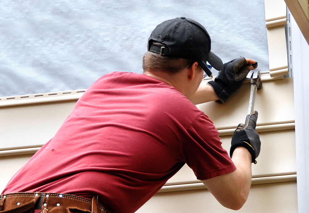Worker in red shirt and tool belt installing siding with a hammer on a house exterior