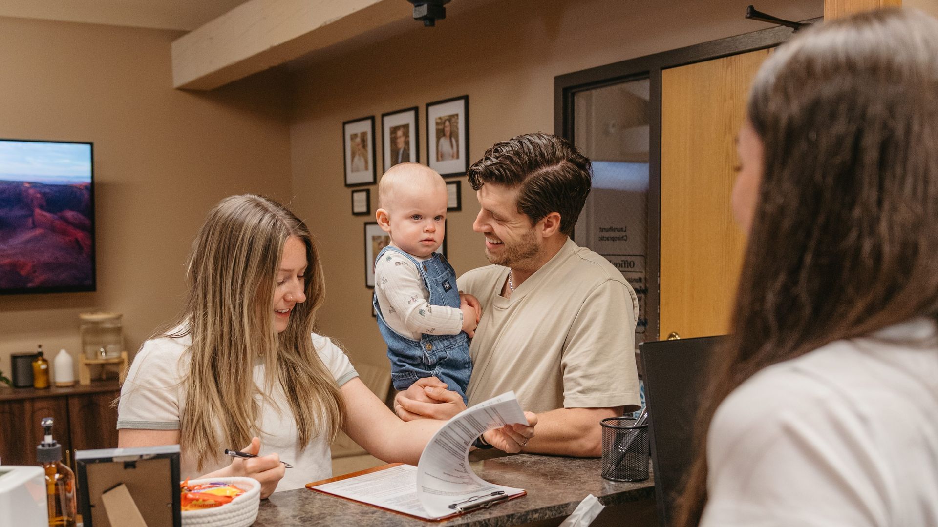 People gathered in a hallway, smiling while one person holds a baby and another looks at papers on a counter.