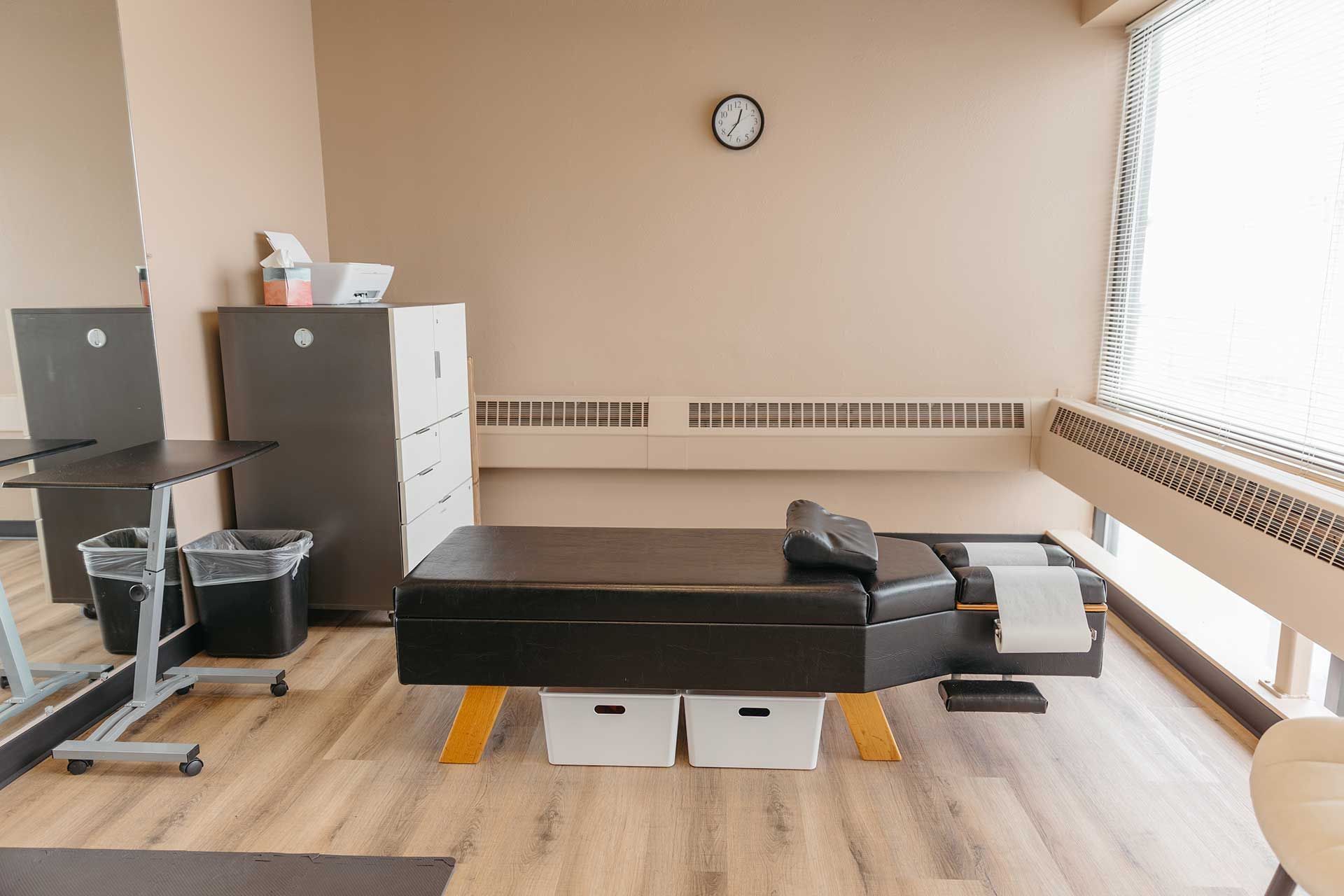 Massage therapy room with black massage table, cabinets, and large windows.