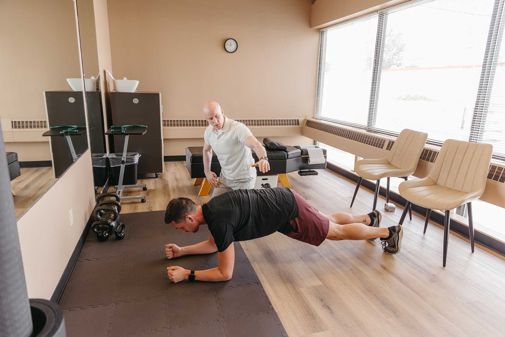 Two people exercising in a bright room, one planking on a mat and one reaching overhead behind him