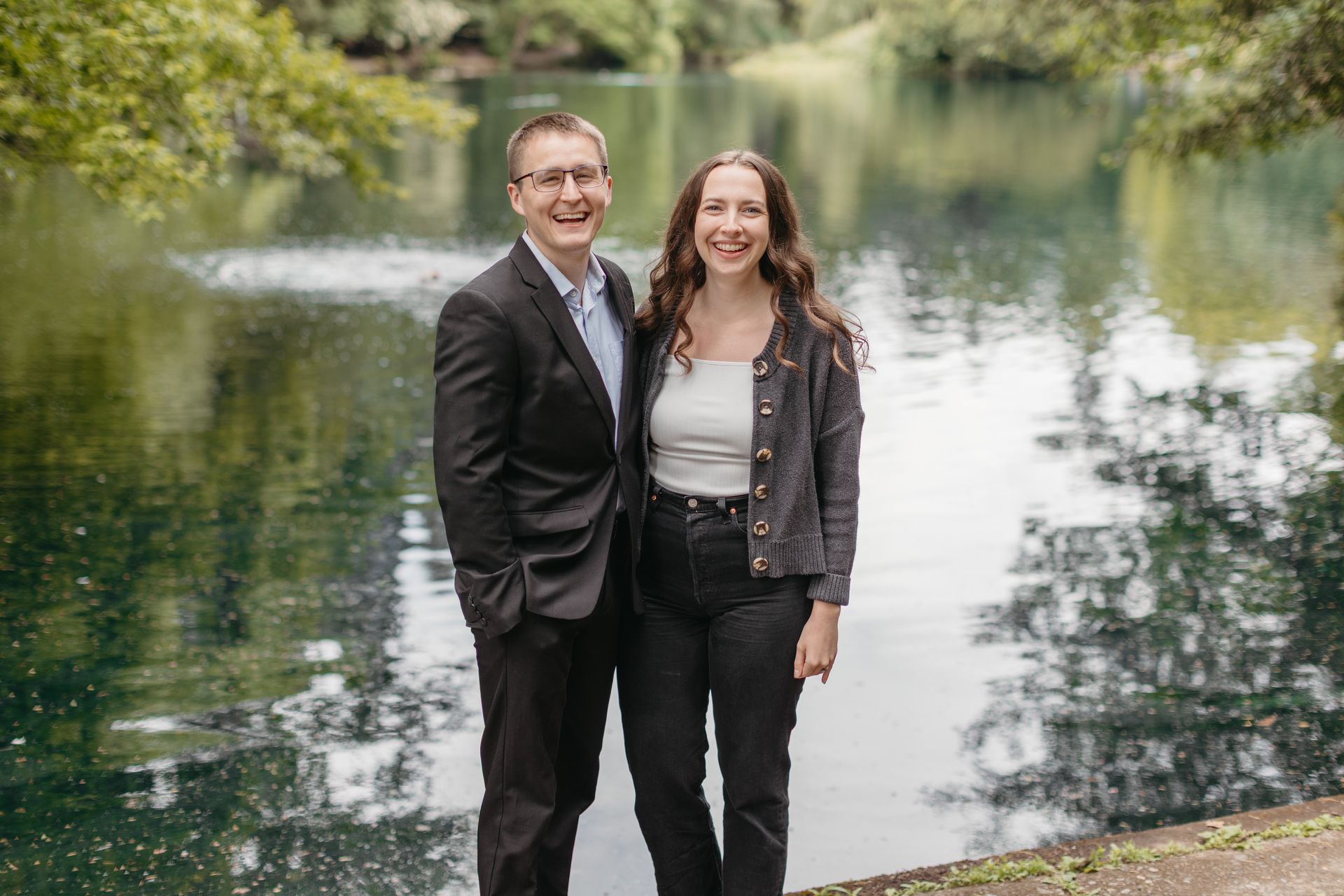 Two people standing by a pond, smiling in a green outdoor park setting