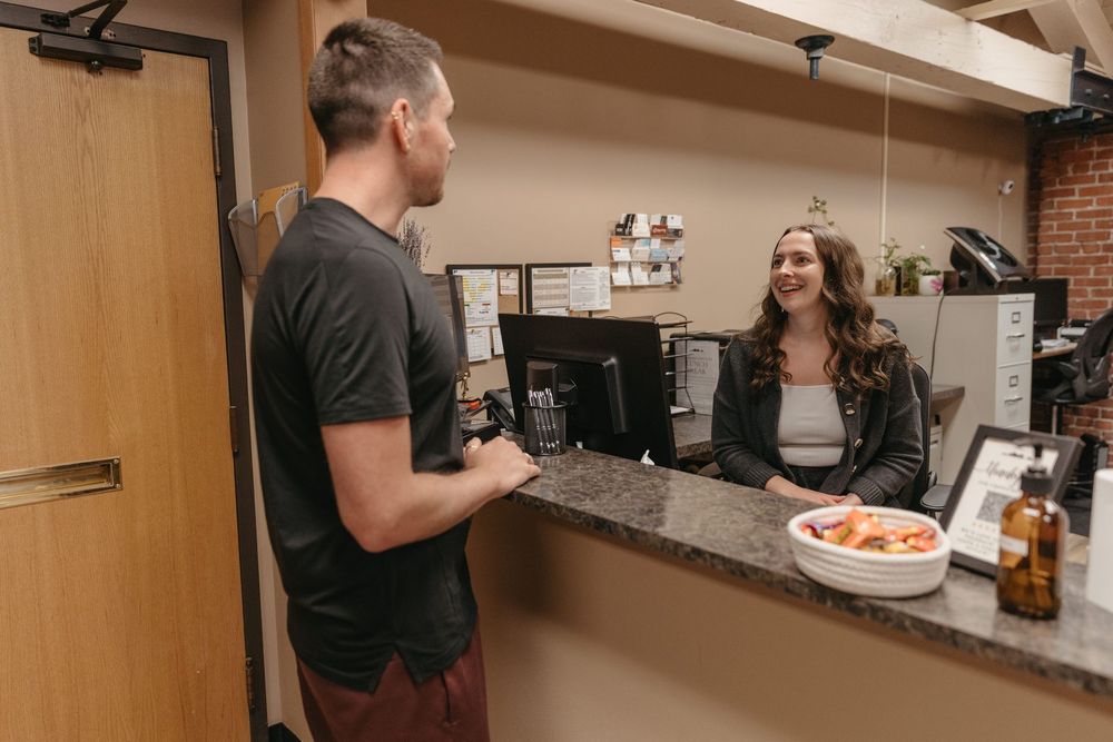 Two people talking at a café counter, with a pastry display and coffee equipment behind the register.