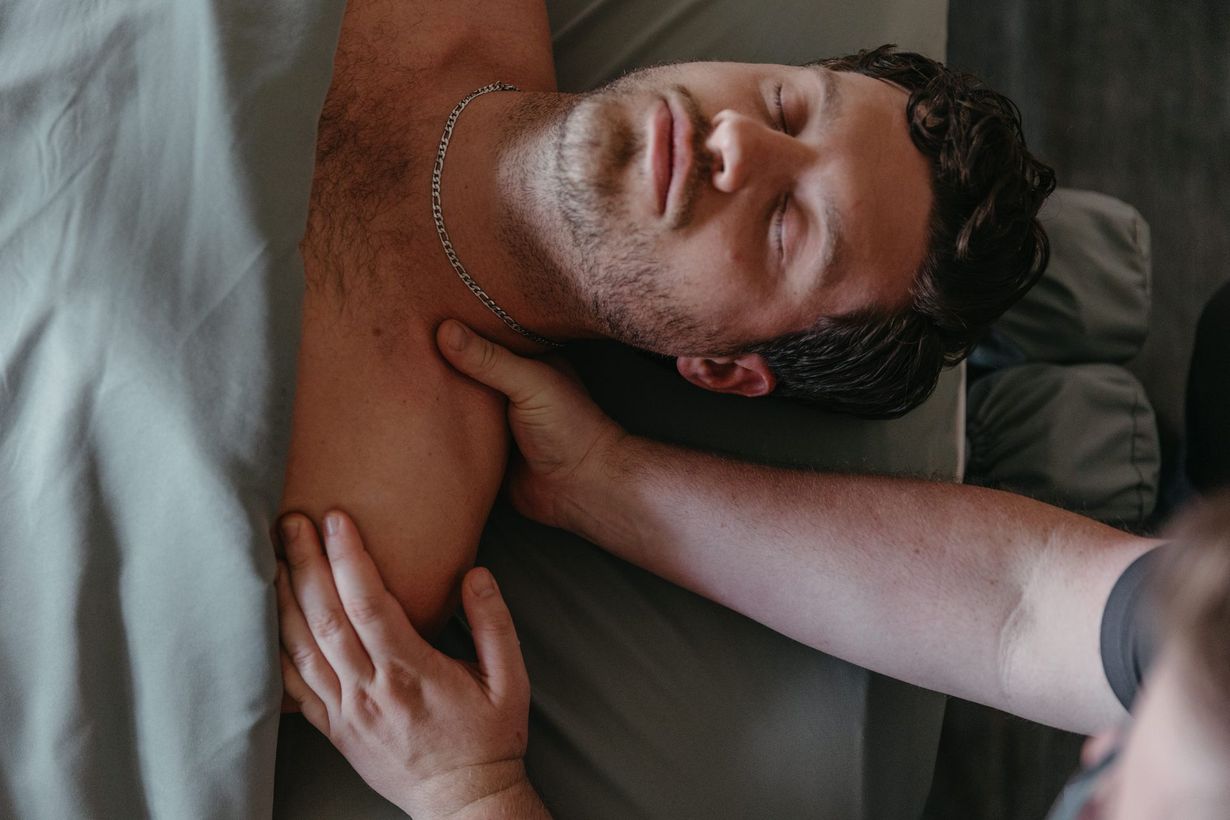 Person lying on a bed, smiling with eyes closed while another person massages their neck and shoulder