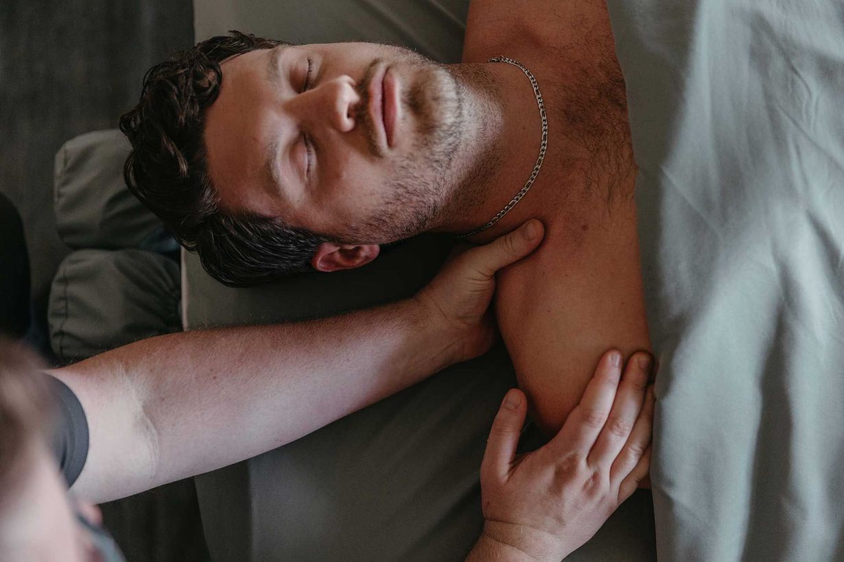 Person lying on a bed, smiling with eyes closed while another person massages their neck and shoulder