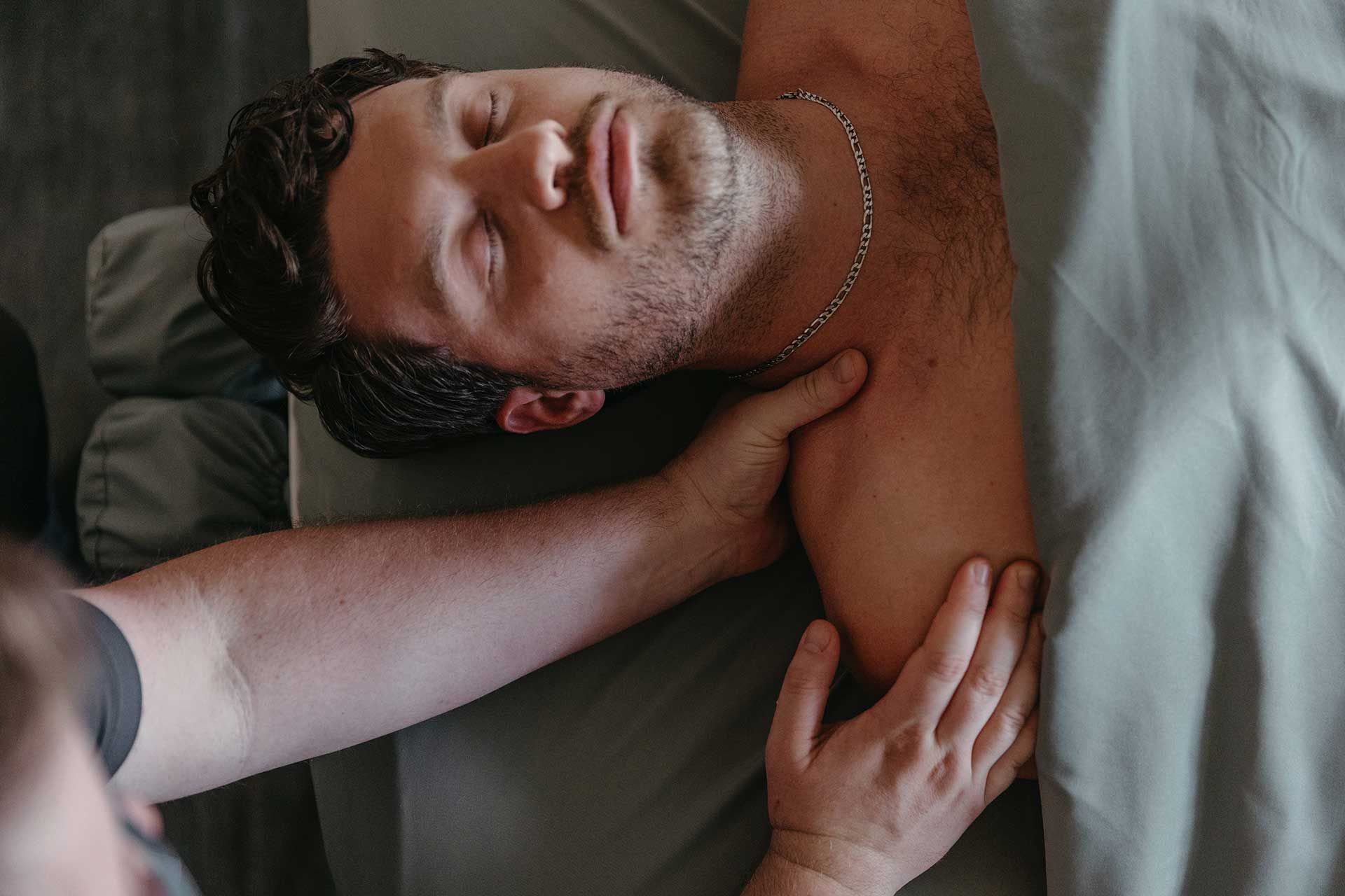 Person lying on a bed, smiling with eyes closed while another person massages their neck and shoulder