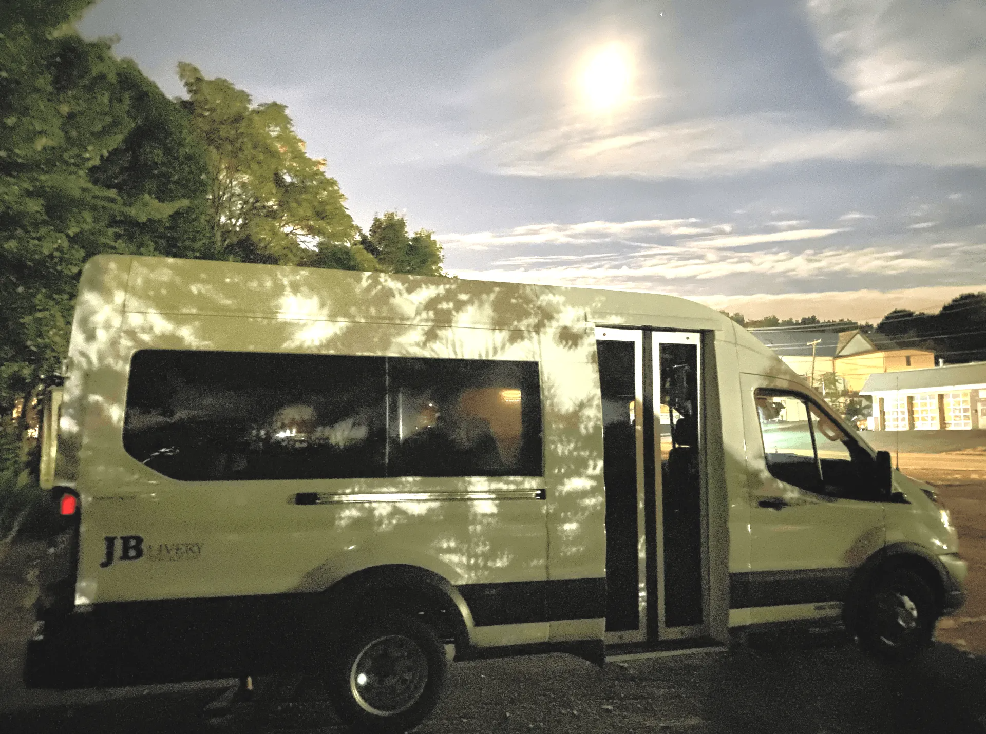 White JB Sport van parked under a partly cloudy sky at dusk. Trees cast shadows on the van.