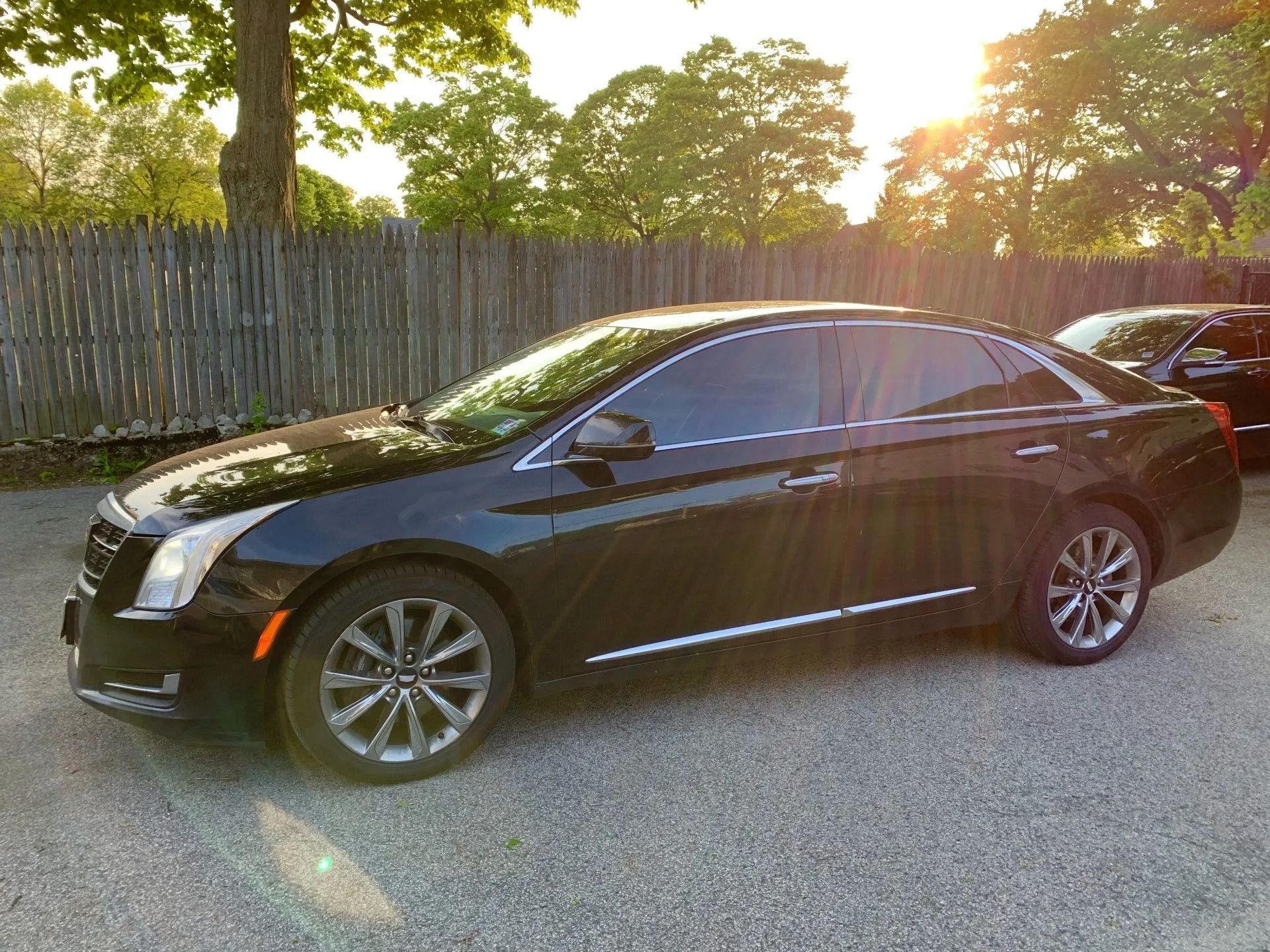 Black Cadillac sedan parked on gravel, against a wooden fence in the evening sun.