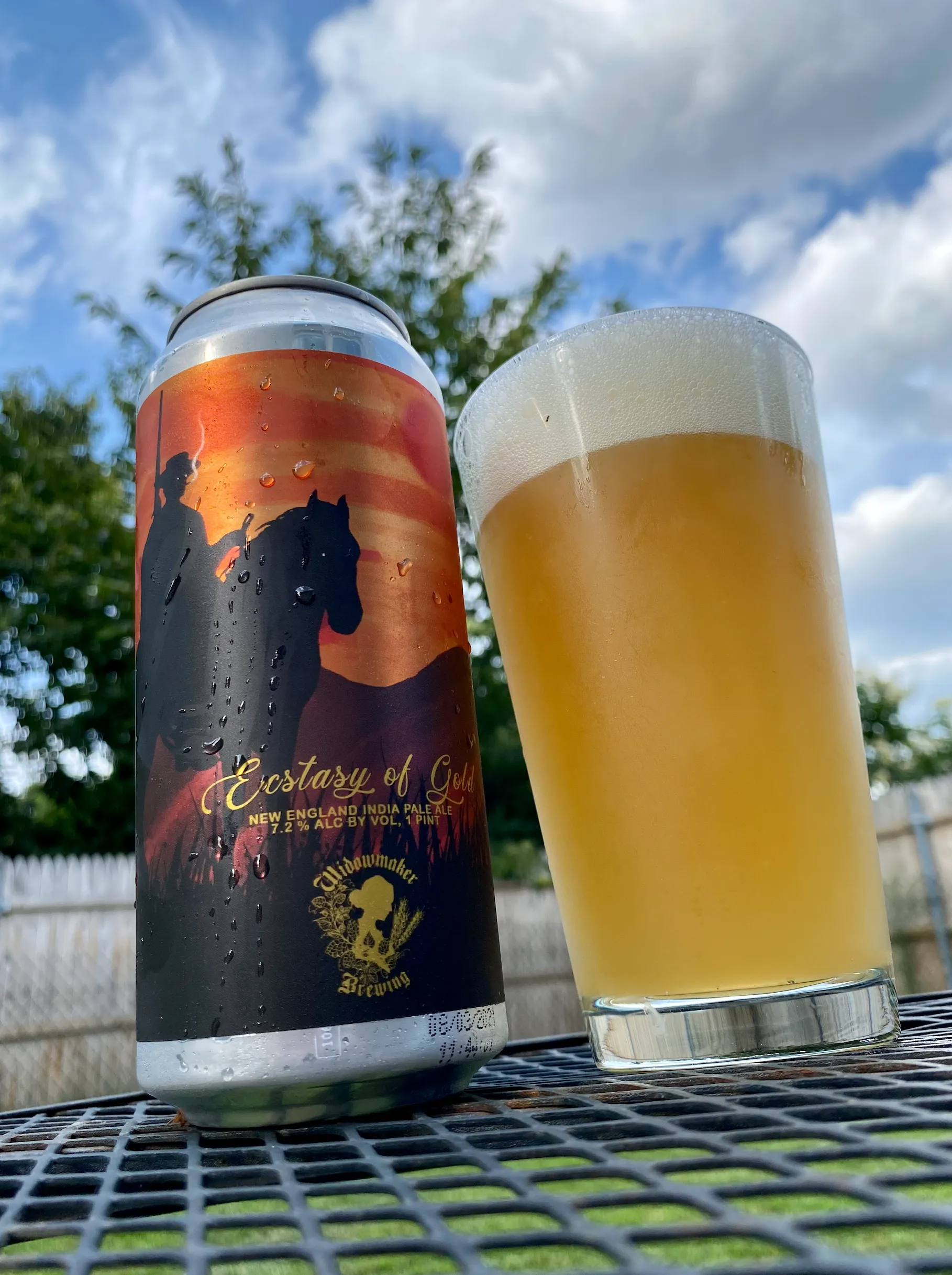 A can of beer and a glass of beer sit on a metal table outside. The can has a silhouette image of a horse and rider.