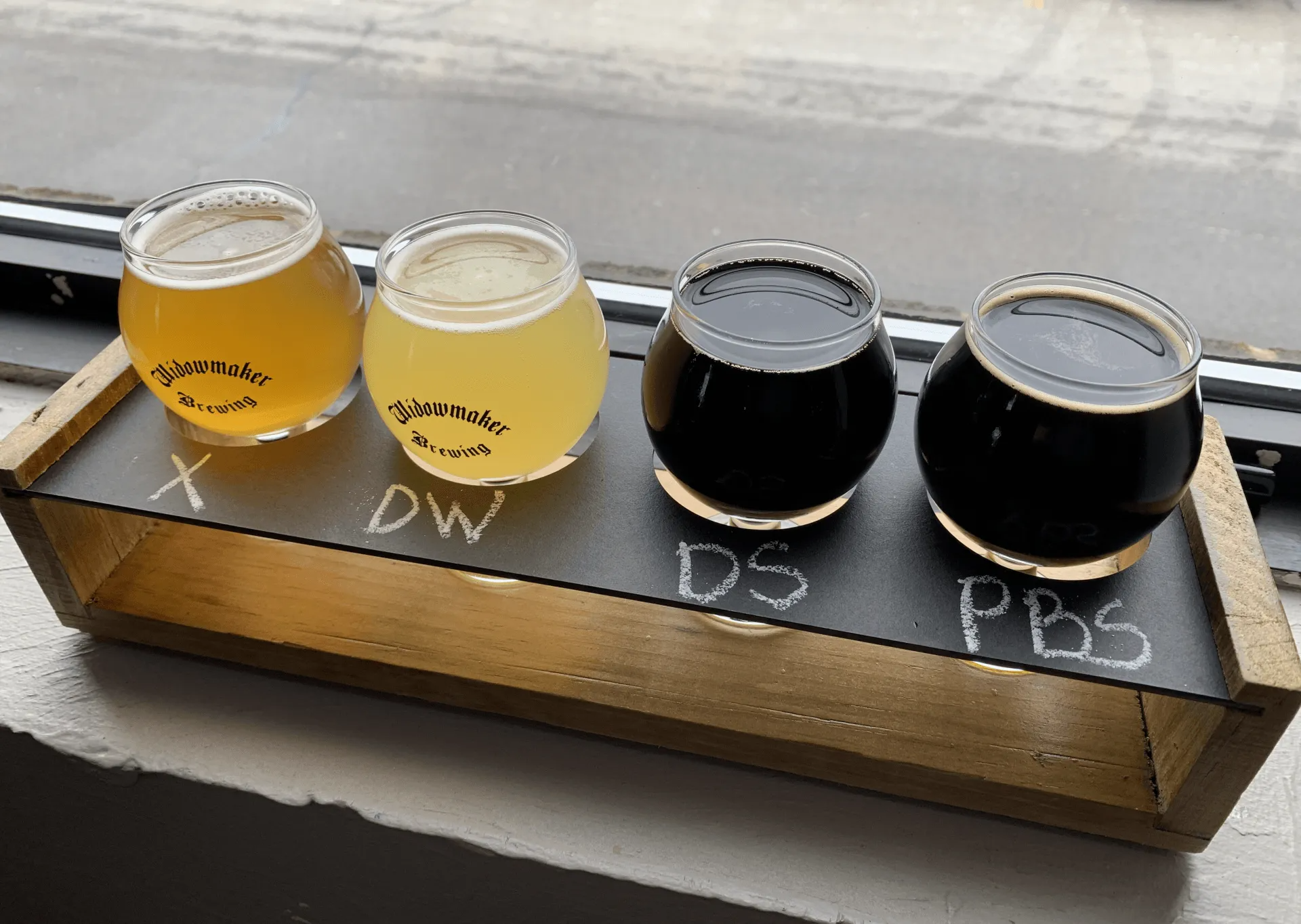 Four beer samples in small glasses on a wooden tray on a windowsill. The beers range in color from light yellow to dark brown.