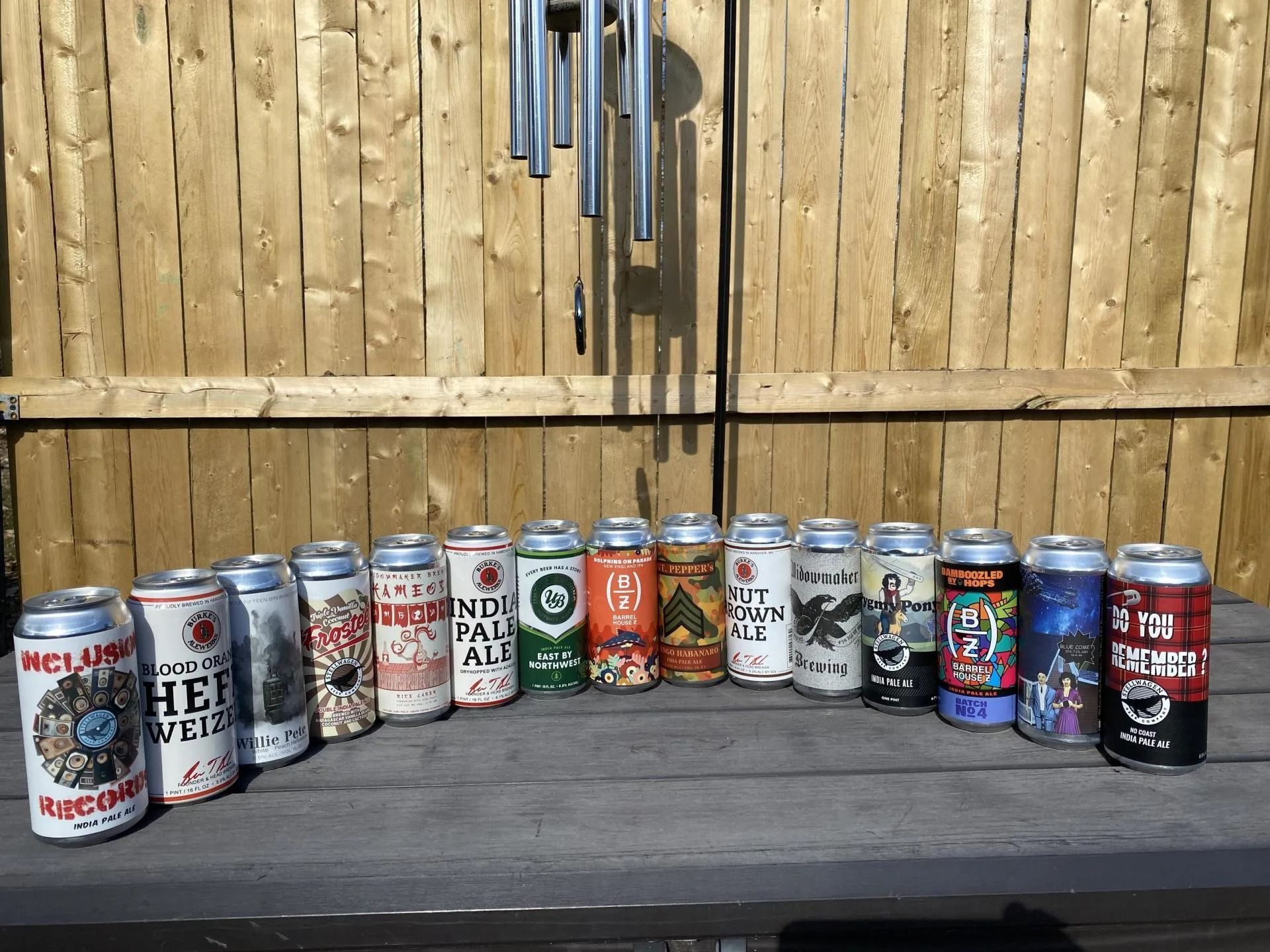 Row of various beer cans on a wooden surface in front of a wood fence and wind chimes.