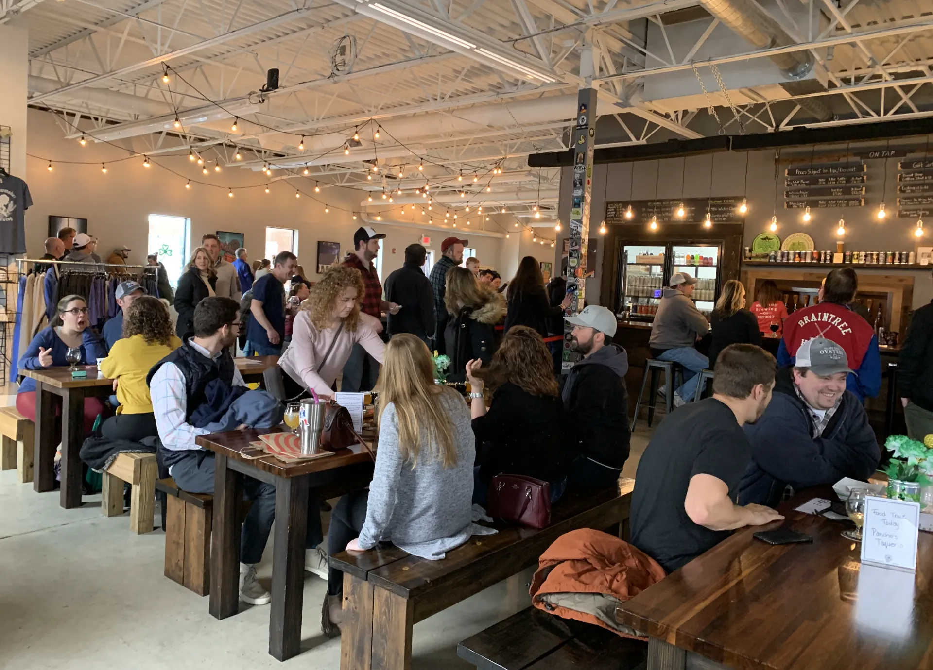 People at tables and standing in a brewery with wooden furniture, string lights, and a bar.