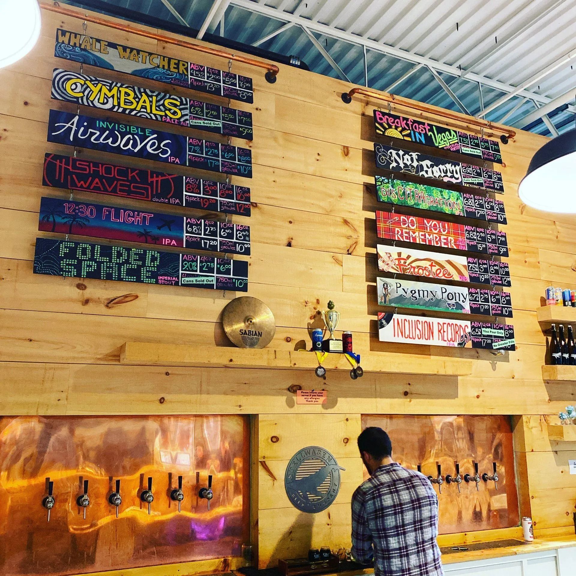 A brewery with wood paneling, copper taps, and colorful beer menu boards. A customer pours beer.