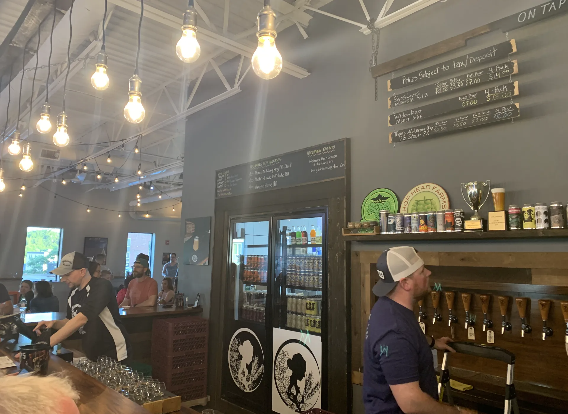Interior of a brewery with patrons, bartenders, beer taps, and chalkboard menus. String lights and awards displayed.