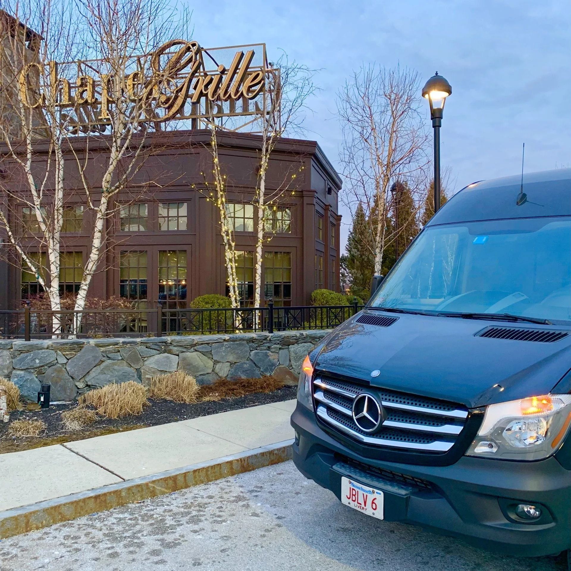 Black Mercedes van parked in front of Chart House Grille restaurant. Dusk setting, stone wall, leafless trees.