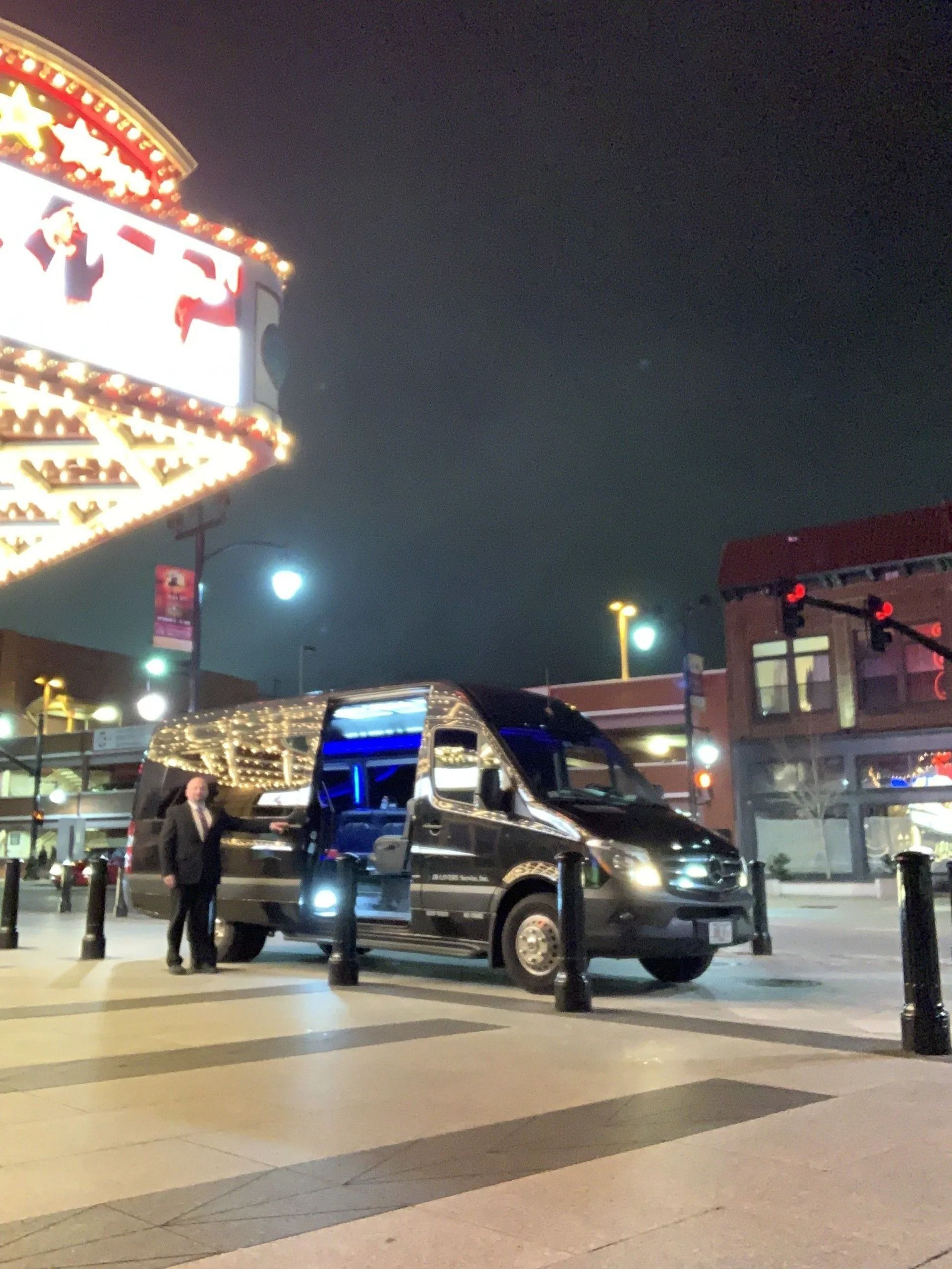 Black van parked at night in front of a lit building, a person in a suit stands near the open door.