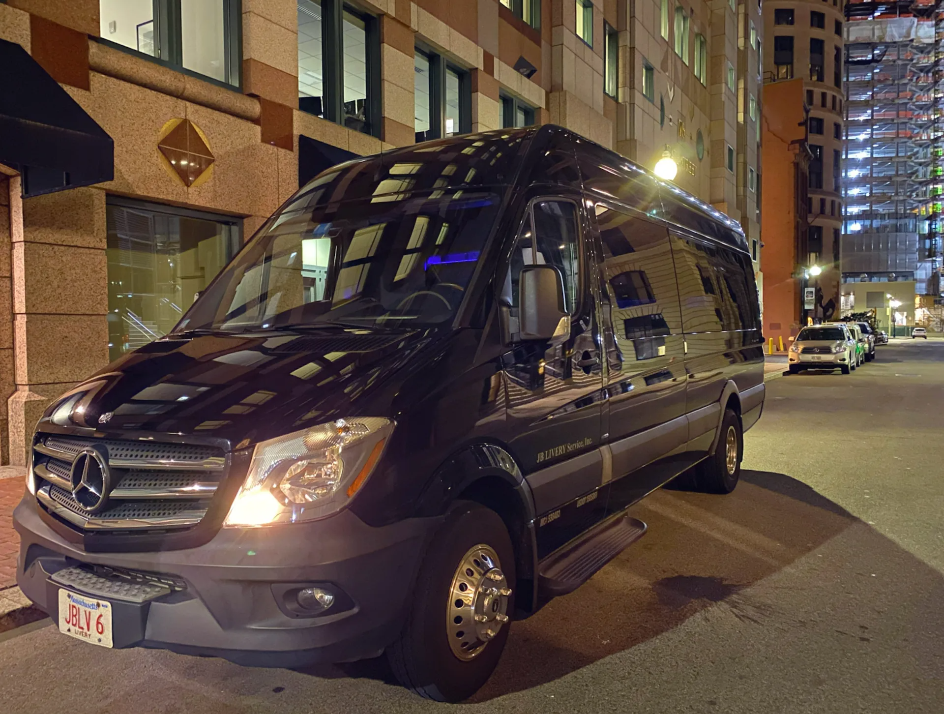 Black Mercedes Sprinter van parked on a city street at night; buildings and streetlights.