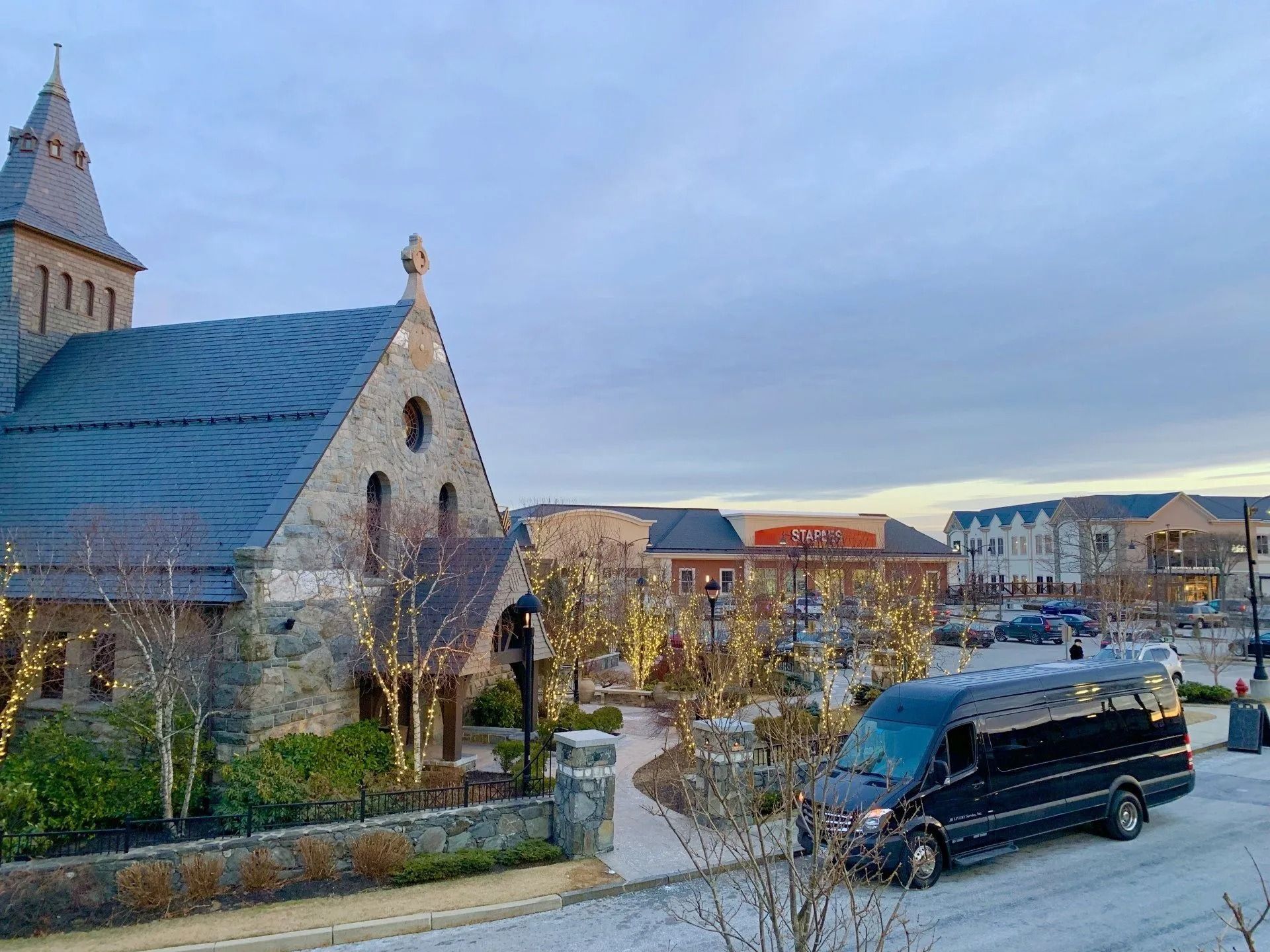 A black van parked on a street in front of a stone church and a shopping area at dusk.