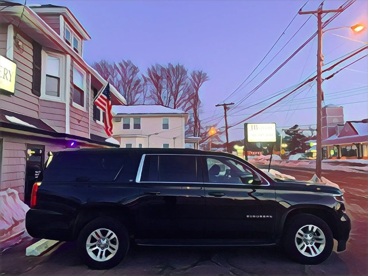 Black Suburban parked on a snowy street next to a building with an American flag. Dusk sky.