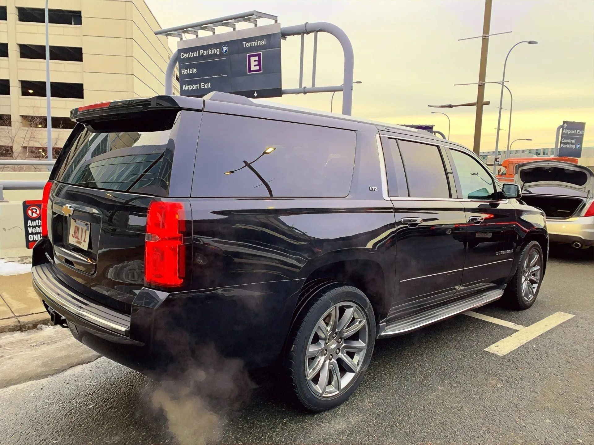 Black SUV parked at an airport terminal, exhaust visible, silver rims, red taillights.