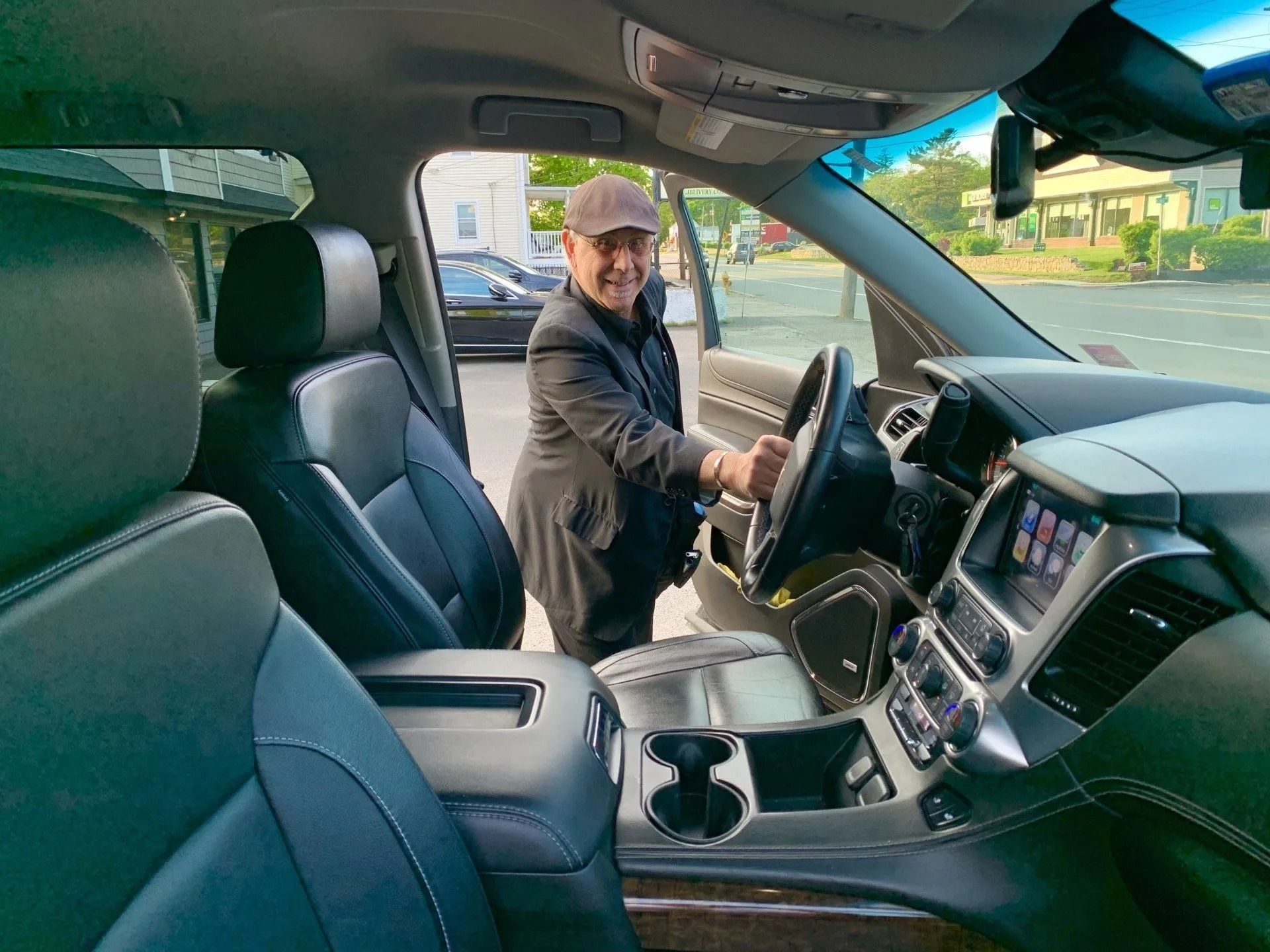 Man in a black jacket and hat exiting a car, reaching for the steering wheel, parked on a street.