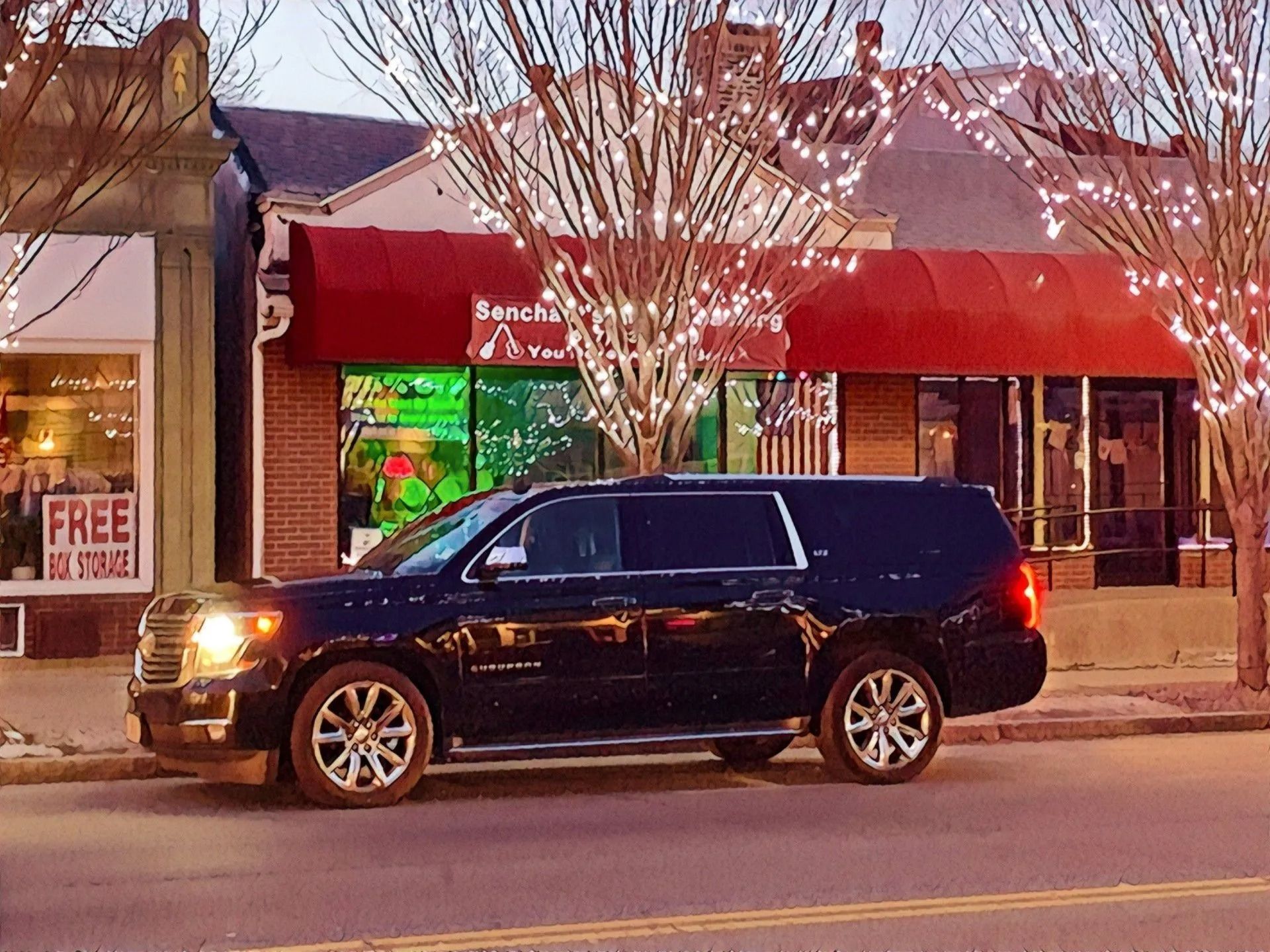 Black SUV parked in front of a brick building with red awning. Christmas lights in the trees.