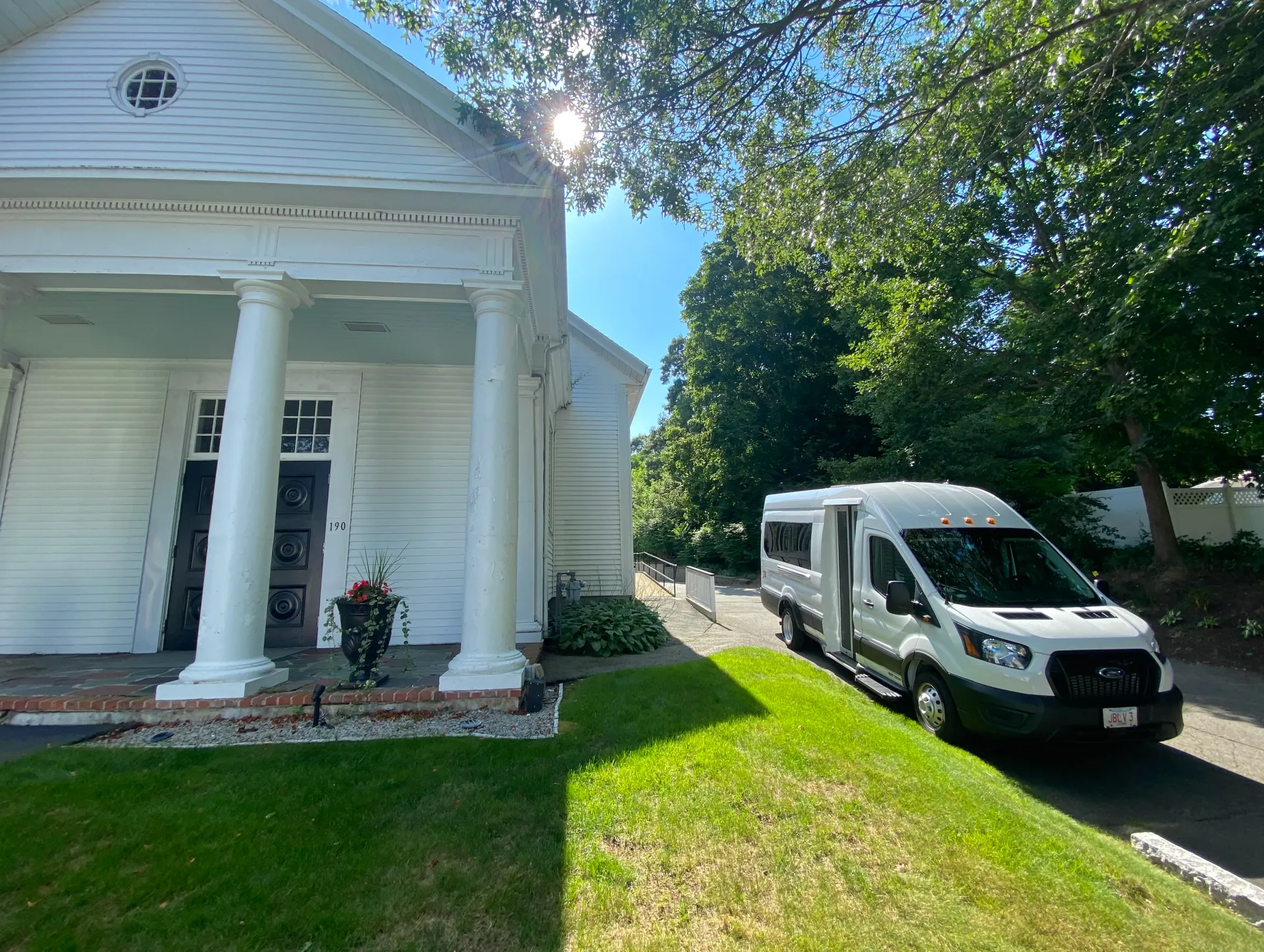 White church with pillars, a white van parked on the right, and green grass on a sunny day.