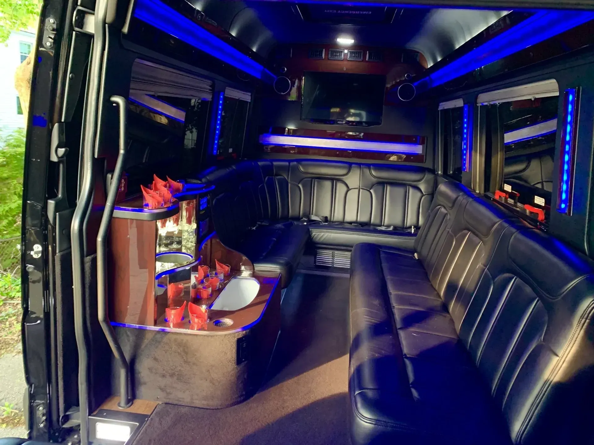 Interior of a black party bus with blue lights, black leather seating, bar, and TV.