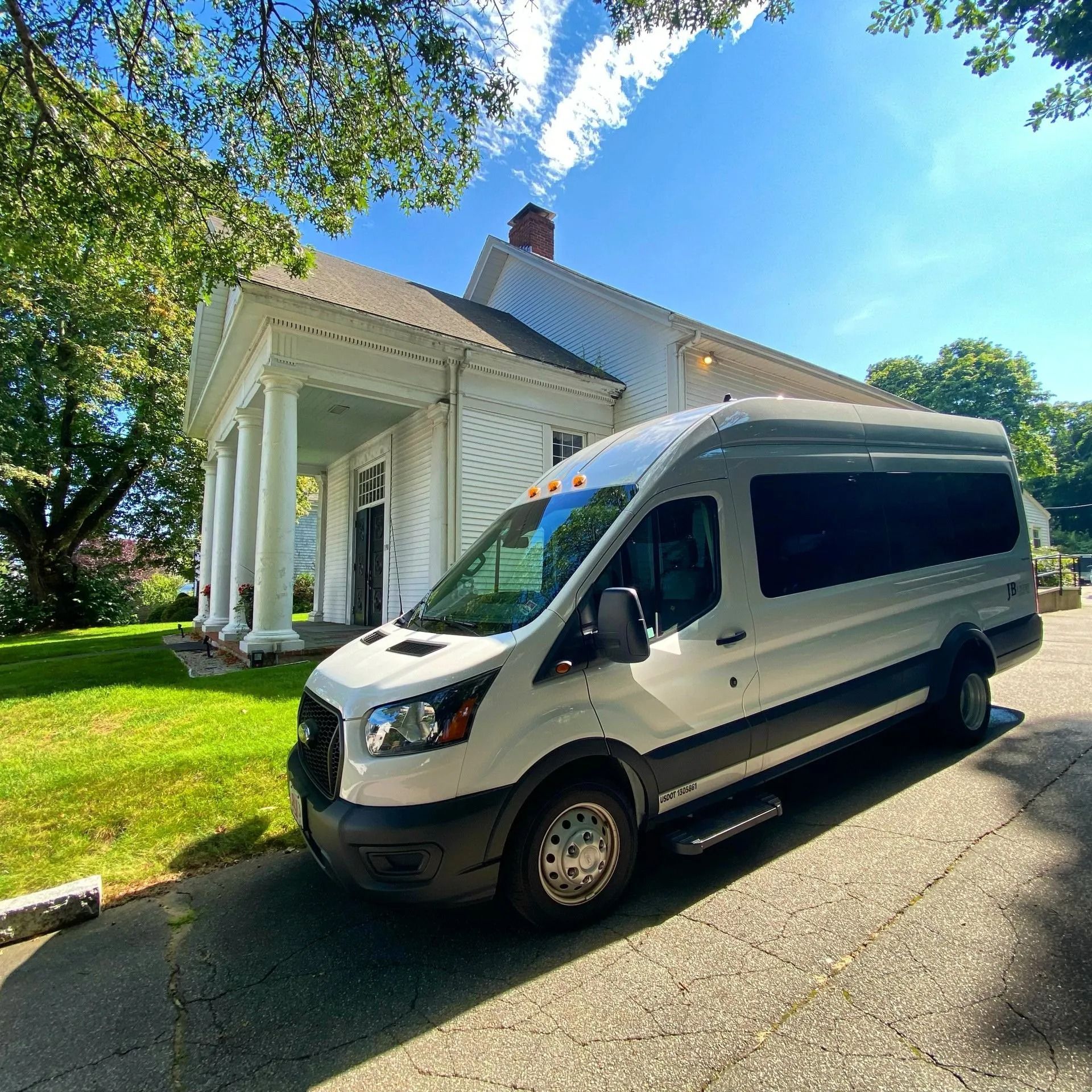White passenger van parked on a driveway in front of a white building with columns; sunny day.