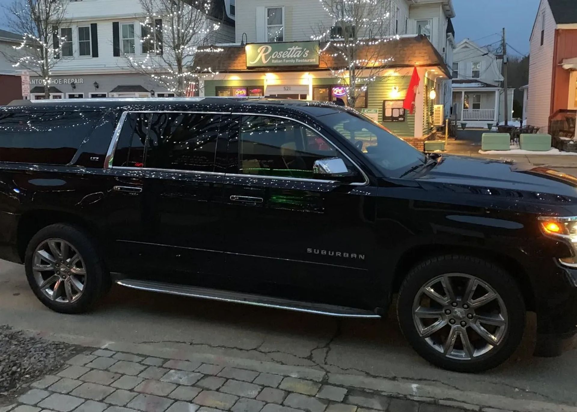 Black SUV parked on a brick street in front of a restaurant with holiday lights.