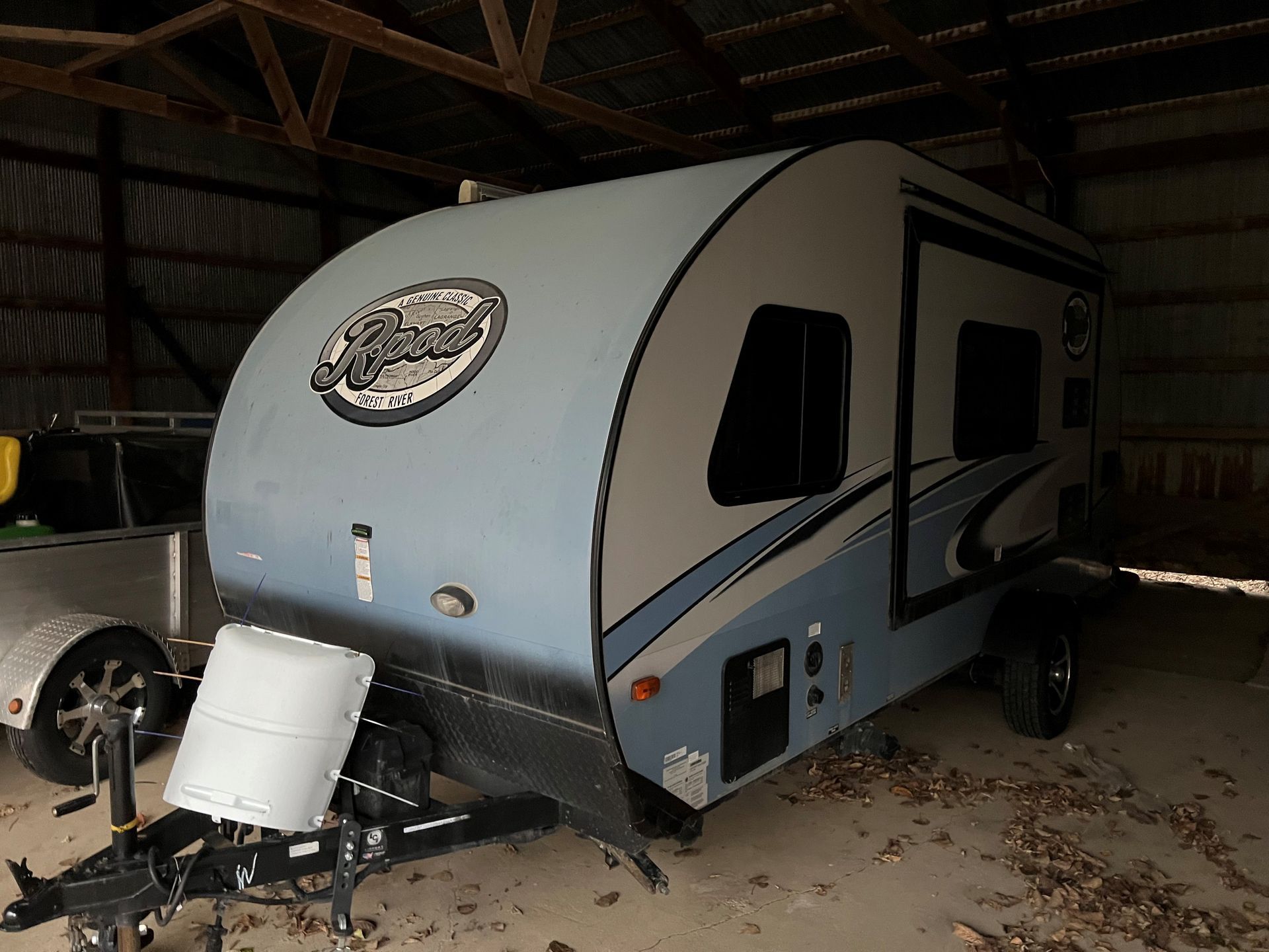 A small blue and white RV trailer is parked in a barn.